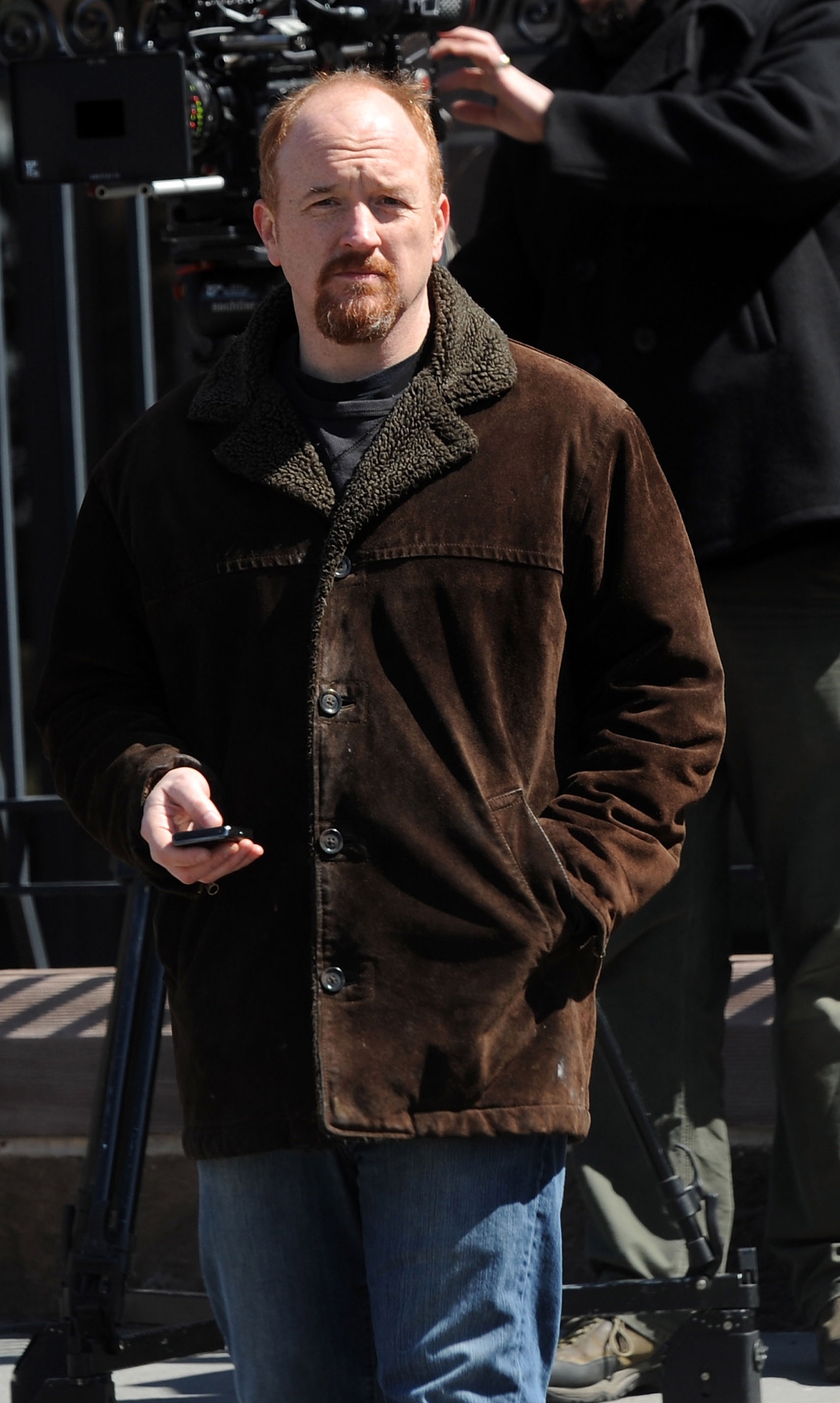 Louis C.K. in casual suede jacket and jeans walks while holding a phone. Film crew with camera visible in the background