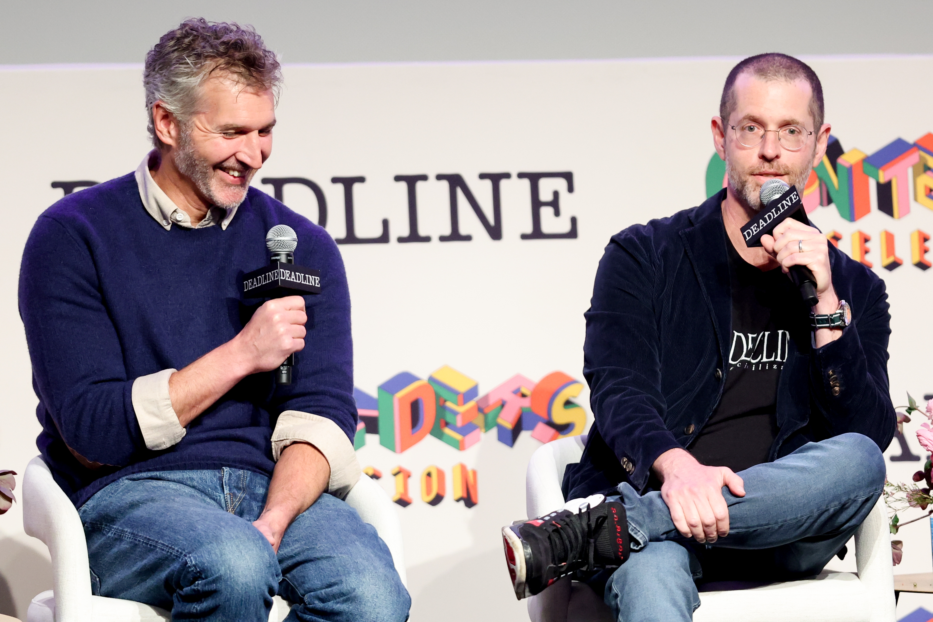 David Benioff and D. B. Weiss sit on stage holding microphones, engaged in a discussion. One wears a sweater and jeans; the other wears a blazer, T-shirt, and jeans