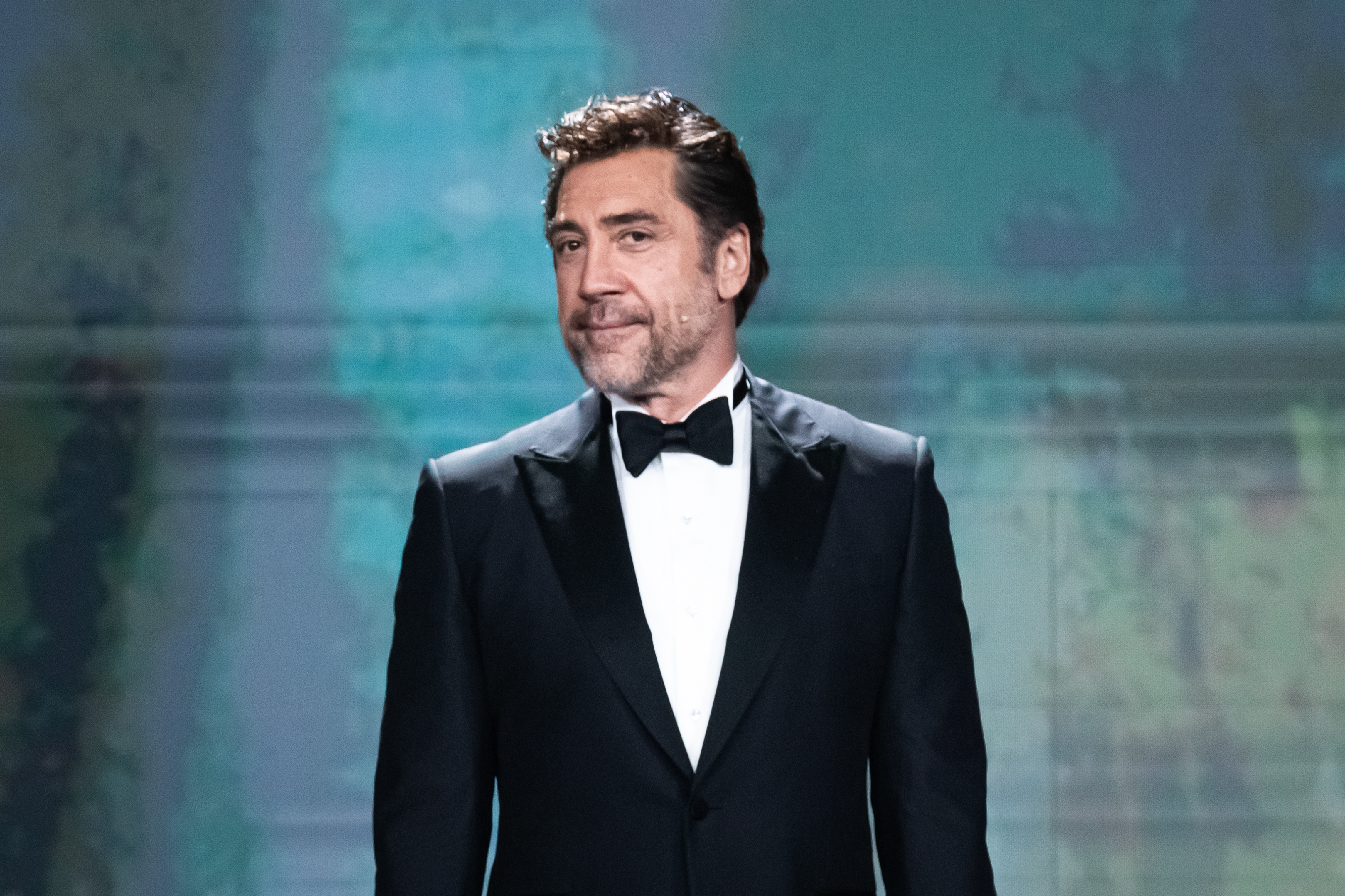Jarier Bardem in a classic tuxedo, smiling slightly, stands on stage against a blurred background