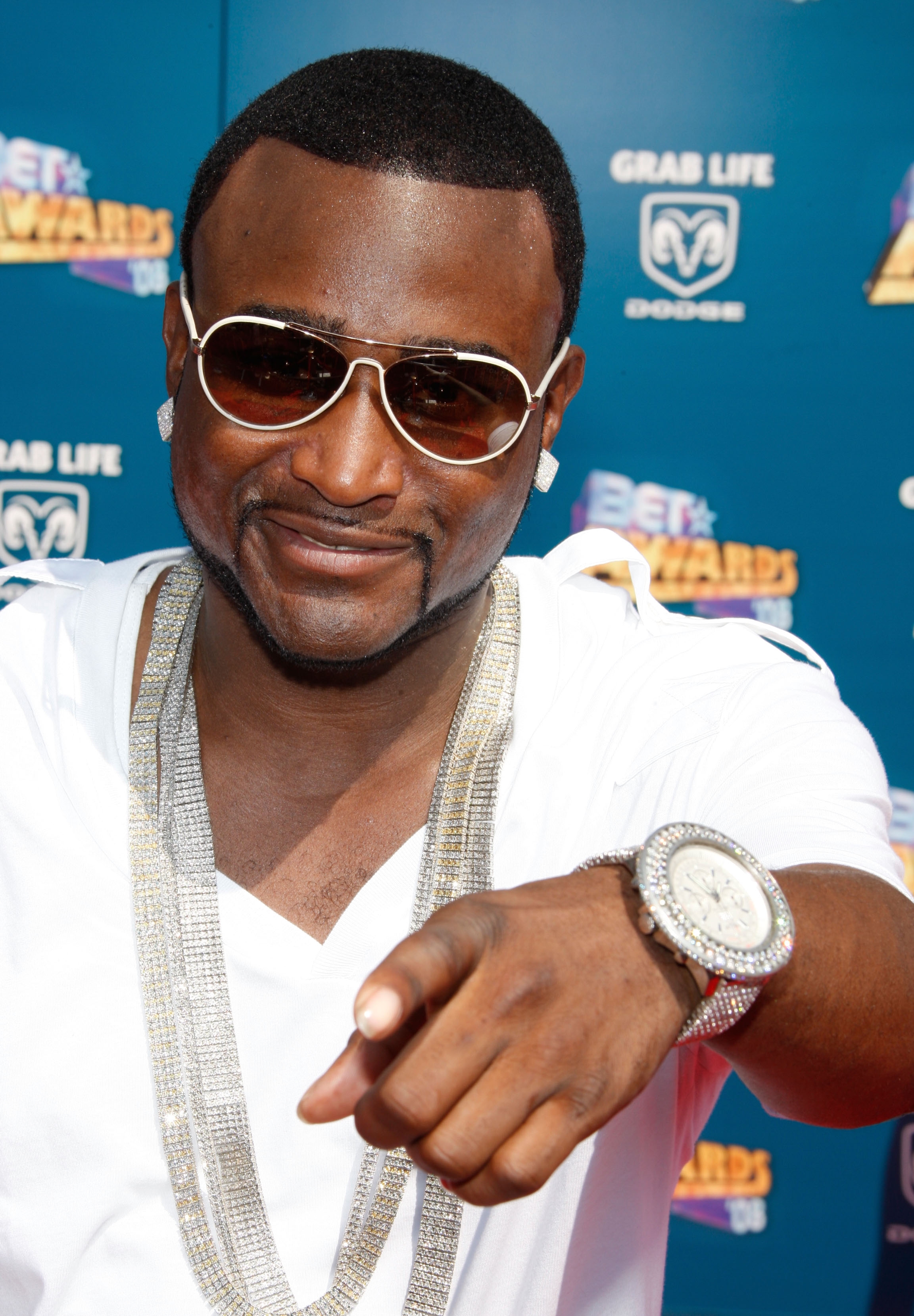 Shawty Lo at an event wearing sunglasses, a white shirt, and large chain necklaces, pointing towards the camera