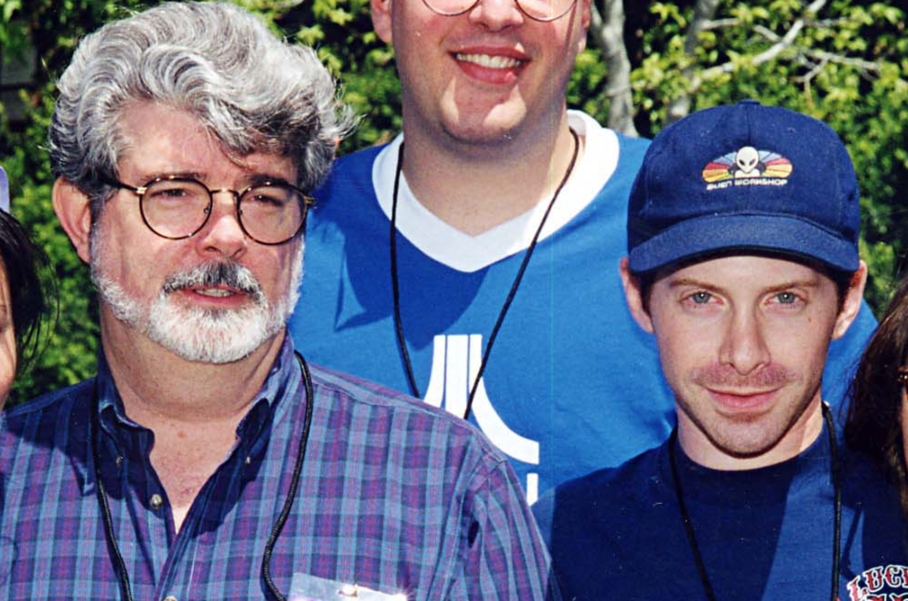 George Lucas and Seth Green pose outdoors, smiling and wearing casual clothes, including a plaid shirt and graphic tees. Trees are visible in the background