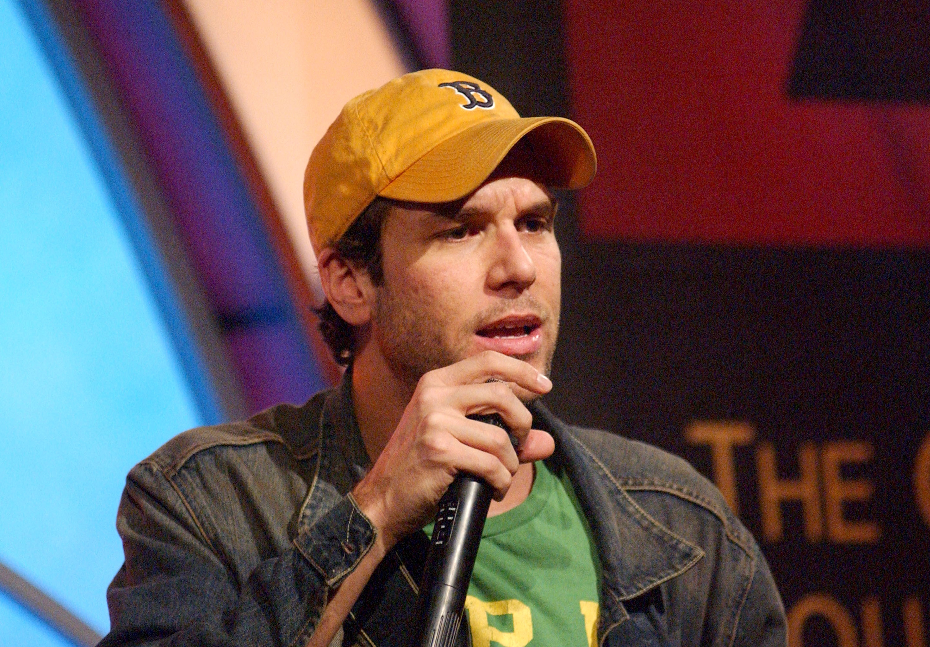Dane COok wearing a denim jacket and a baseball cap, holding a microphone on stage during a performance