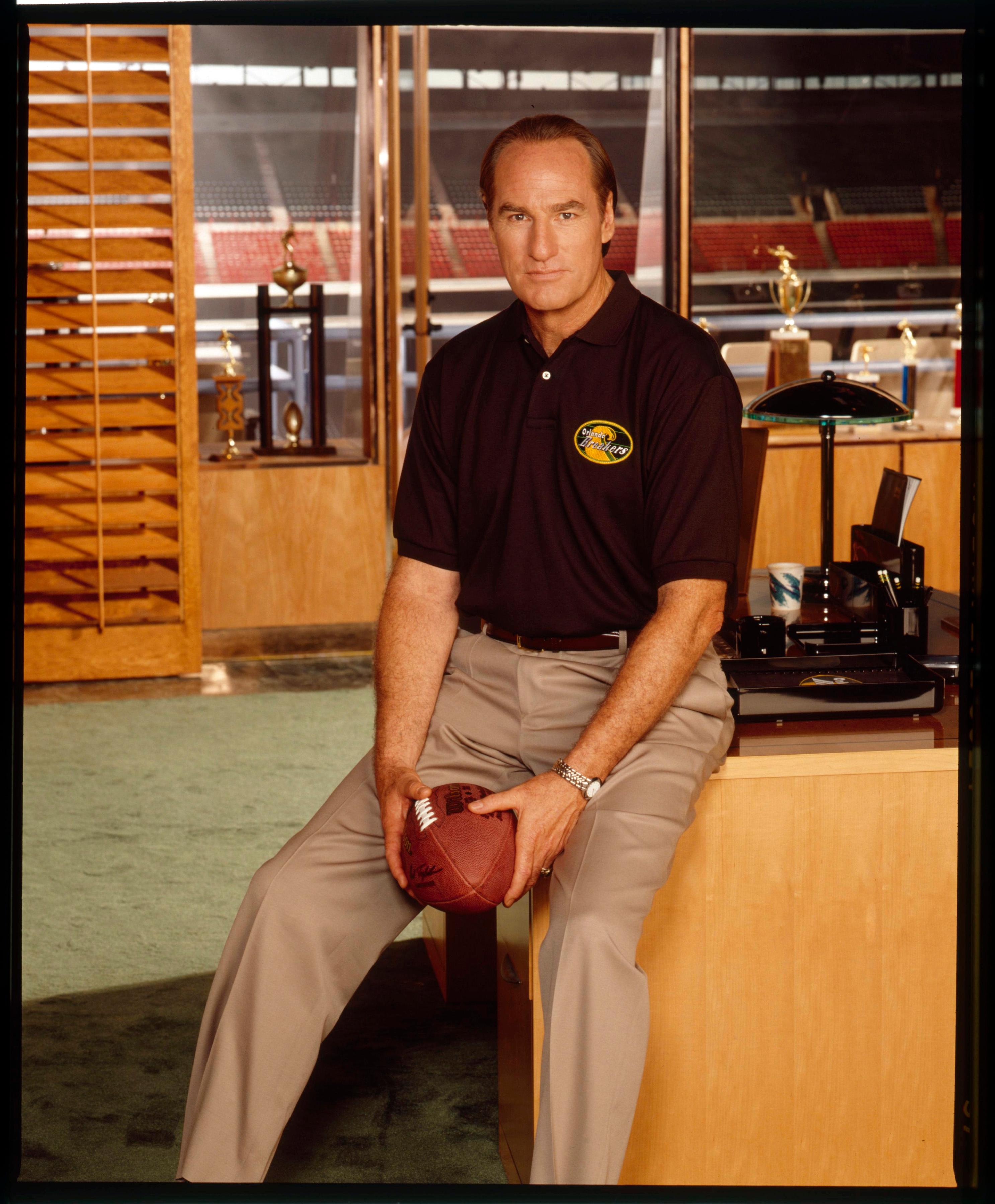 Craig T Nelson sitting on a desk holding a football in an office with stadium view. Wearing a polo shirt and khaki pants. Sports memorabilia in the background