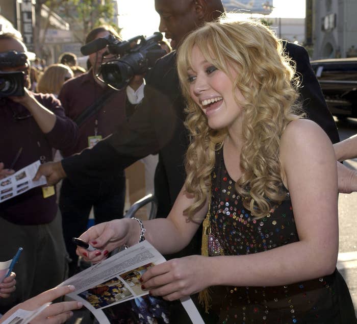 Hilary Duff in sequined gown signing autographs while smiling, surrounded by photographers and fans at an event
