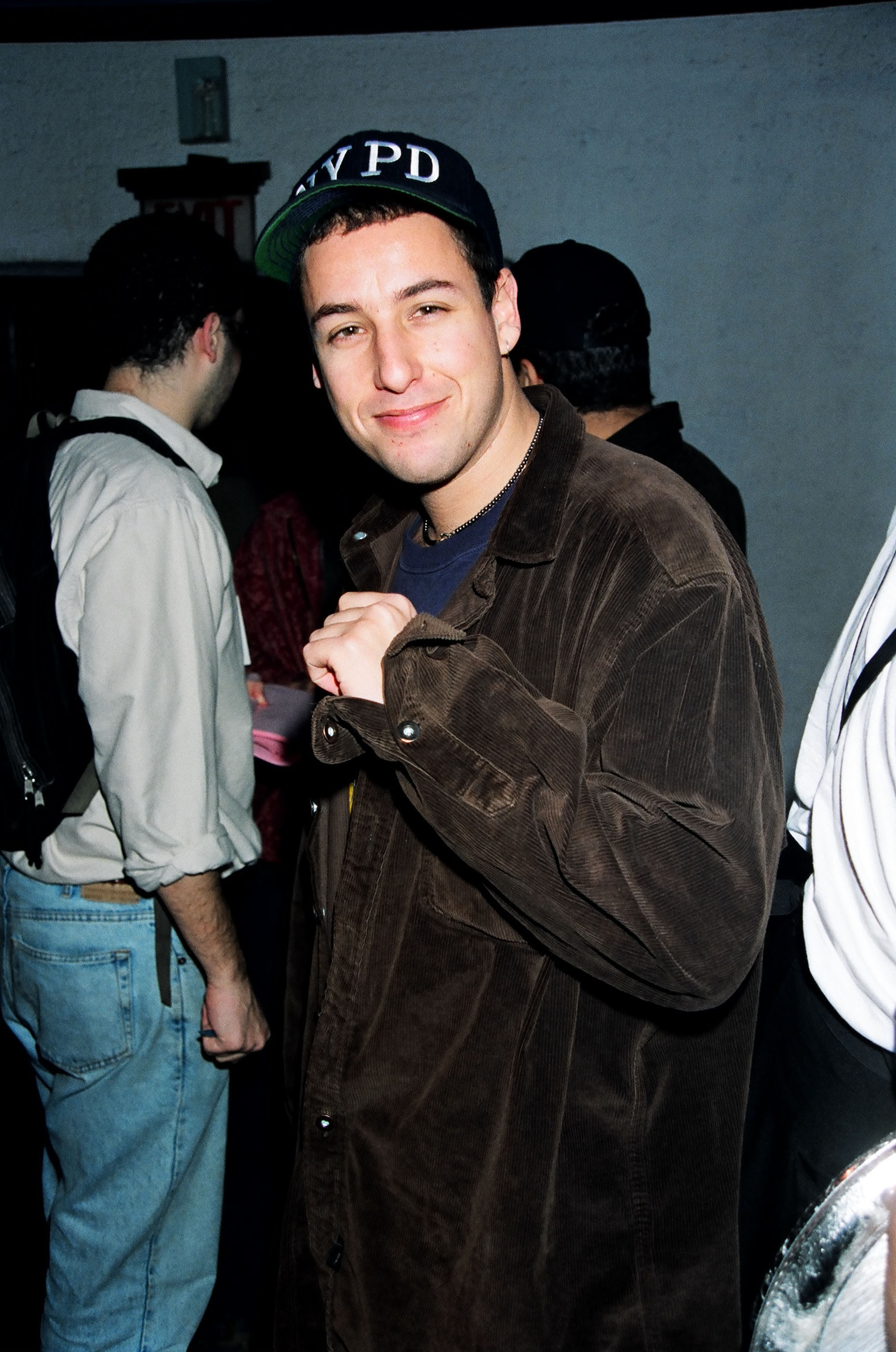 Adam Sandler wearing an NYPD cap and casual jacket at an informal event, smiling at the camera