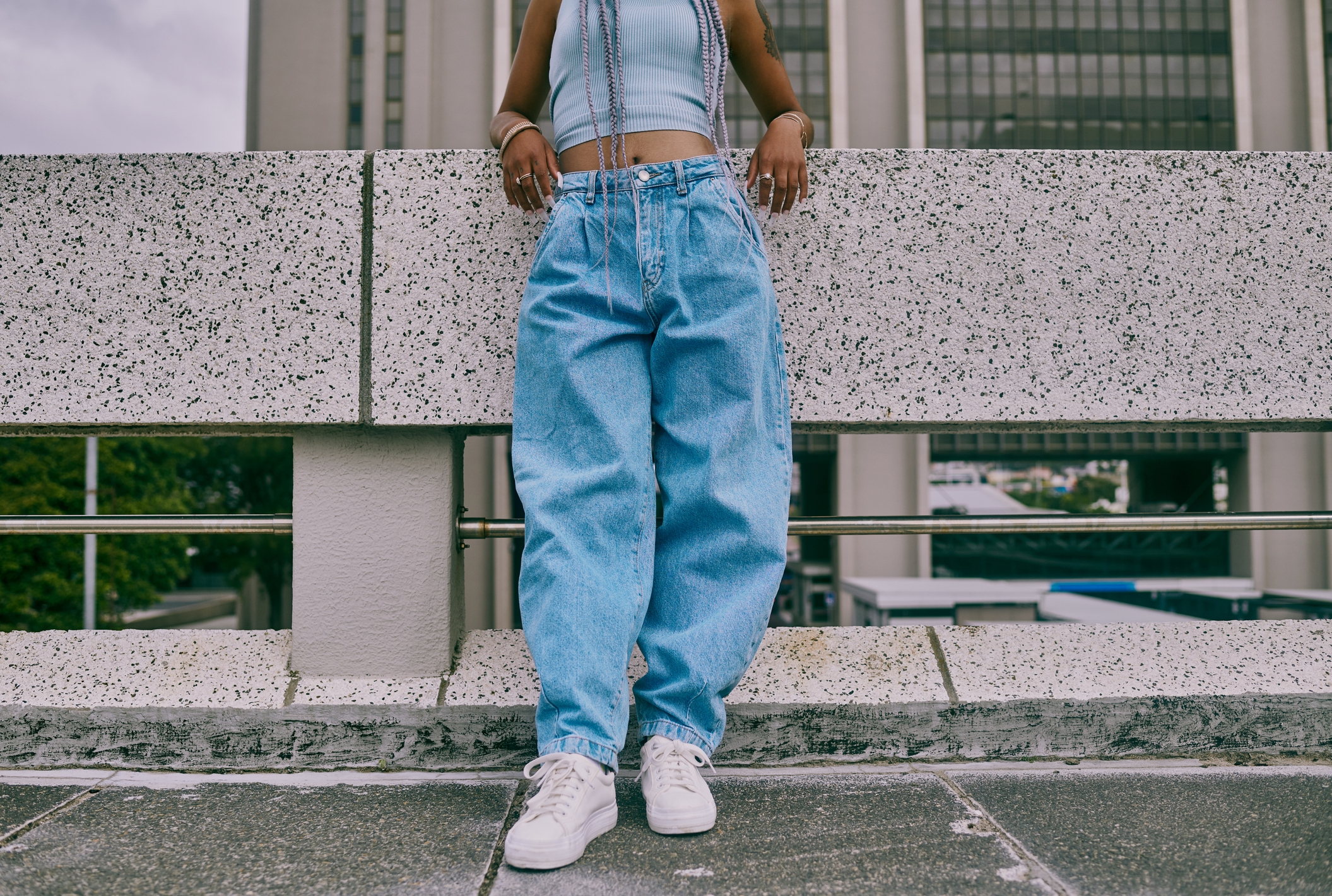 Person i avlingstopp og baggy jeans lener seg mot et rekkverk i urbane omgivelser. Ansiktet deres er ikke synlig