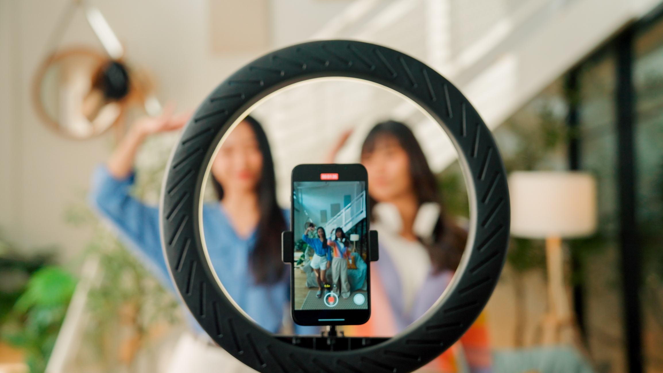 To personer tar en selfie gjennom et ringlys, med en smarttelefon som fanger bildet. De er tilfeldig kledd og i et moderne, godt opplyst rom