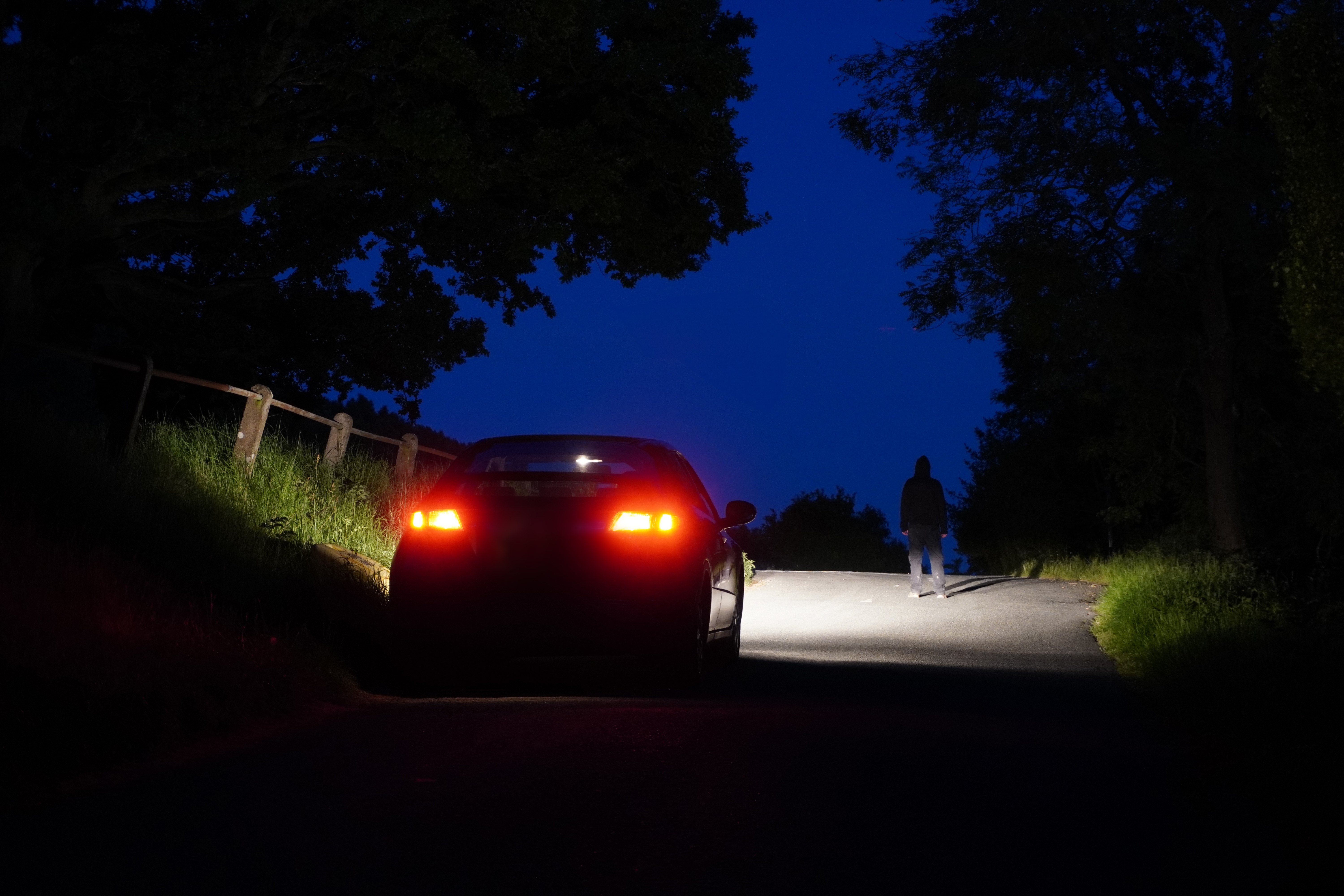 A car is parked on a dimly lit road while a person walks ahead illuminated by headlights