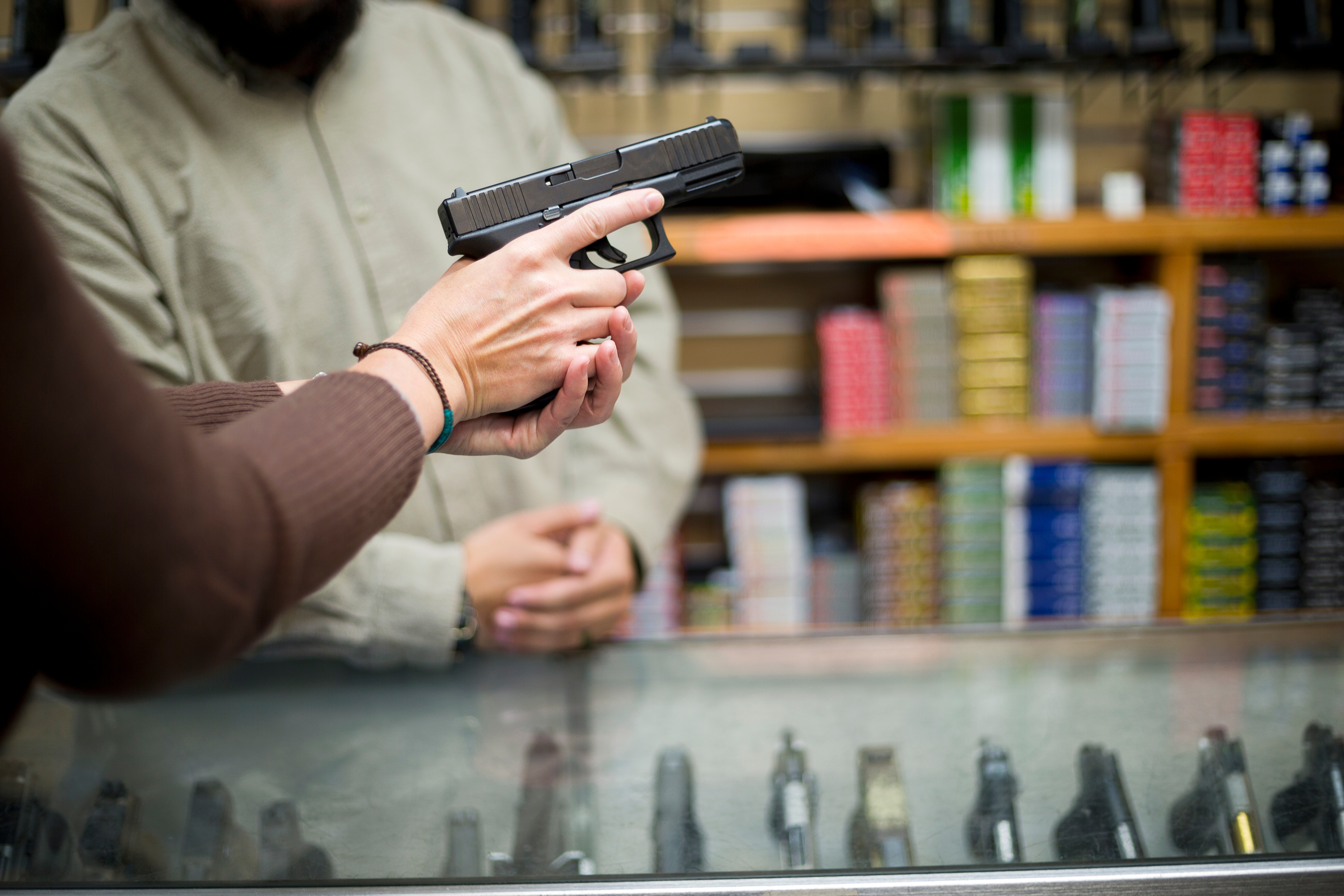 Person holding a handgun at a store counter with another person in the background, showcasing a selection of firearms