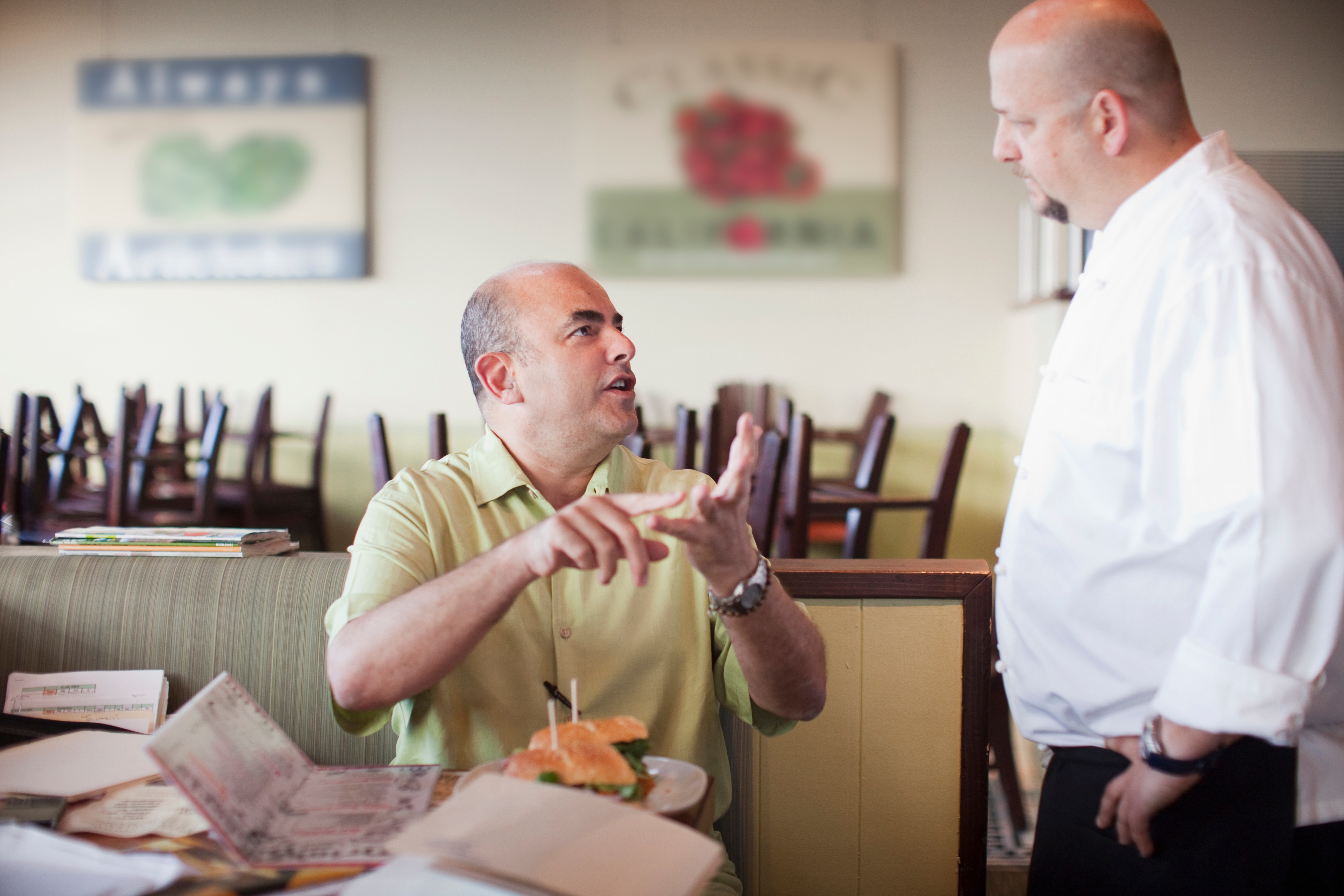 Two people in a restaurant are engaged in discussion. One is seated, gesturing, with documents on the table. A chef stands nearby attentively