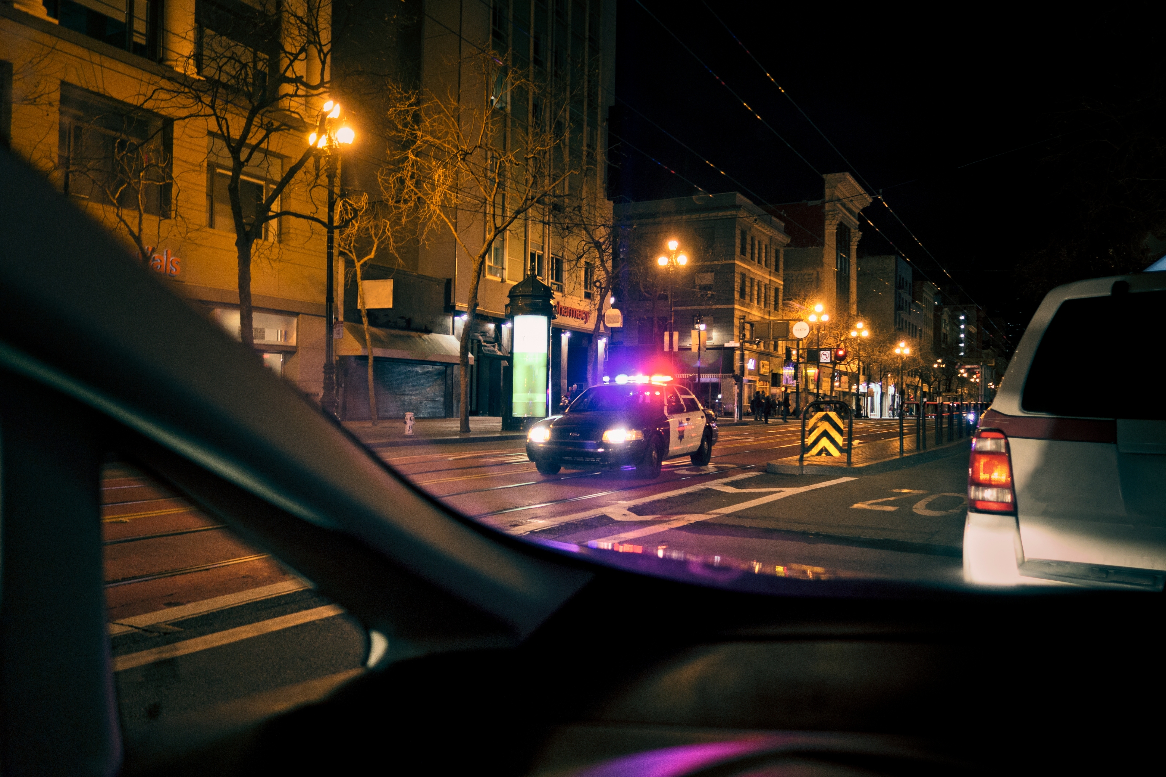 Police car with flashing lights blocks a city street at night, viewed from inside another vehicle