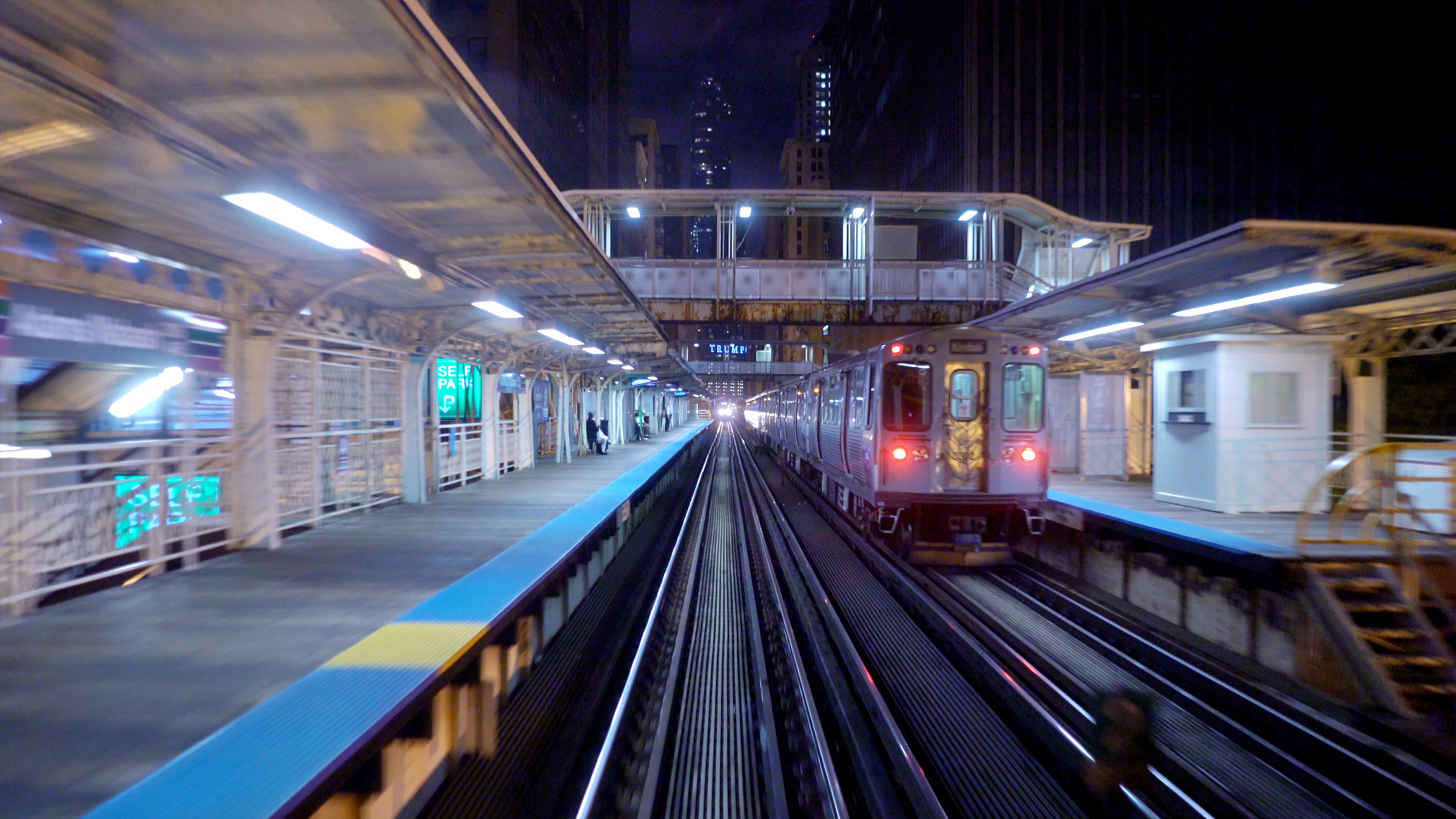 An urban train moves swiftly through an elevated city station at night with blurred lights and skyscrapers in the background
