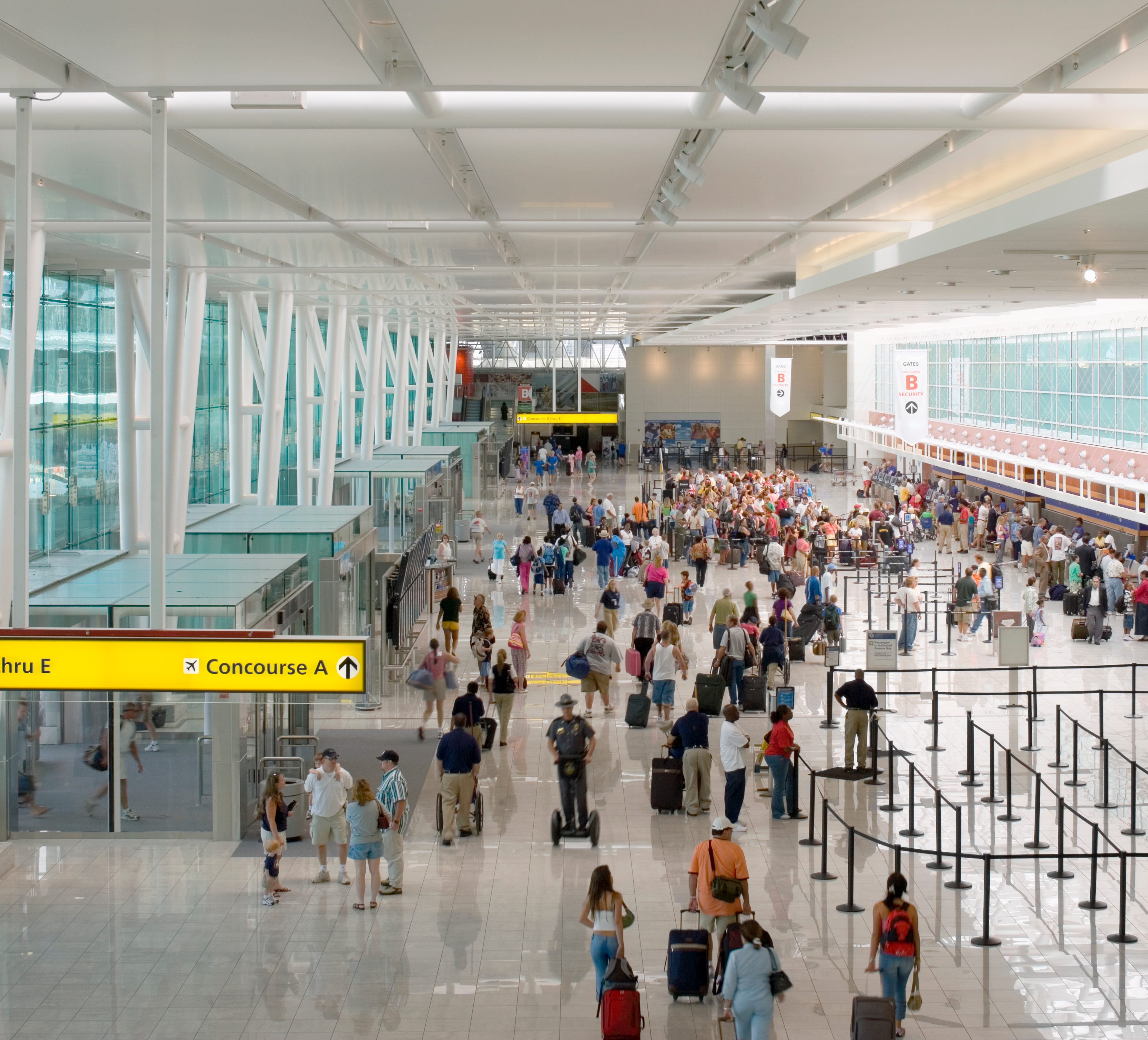 Busy airport terminal with travelers walking with luggage, lined up at check-in counters, and signs directing to gates and concourses