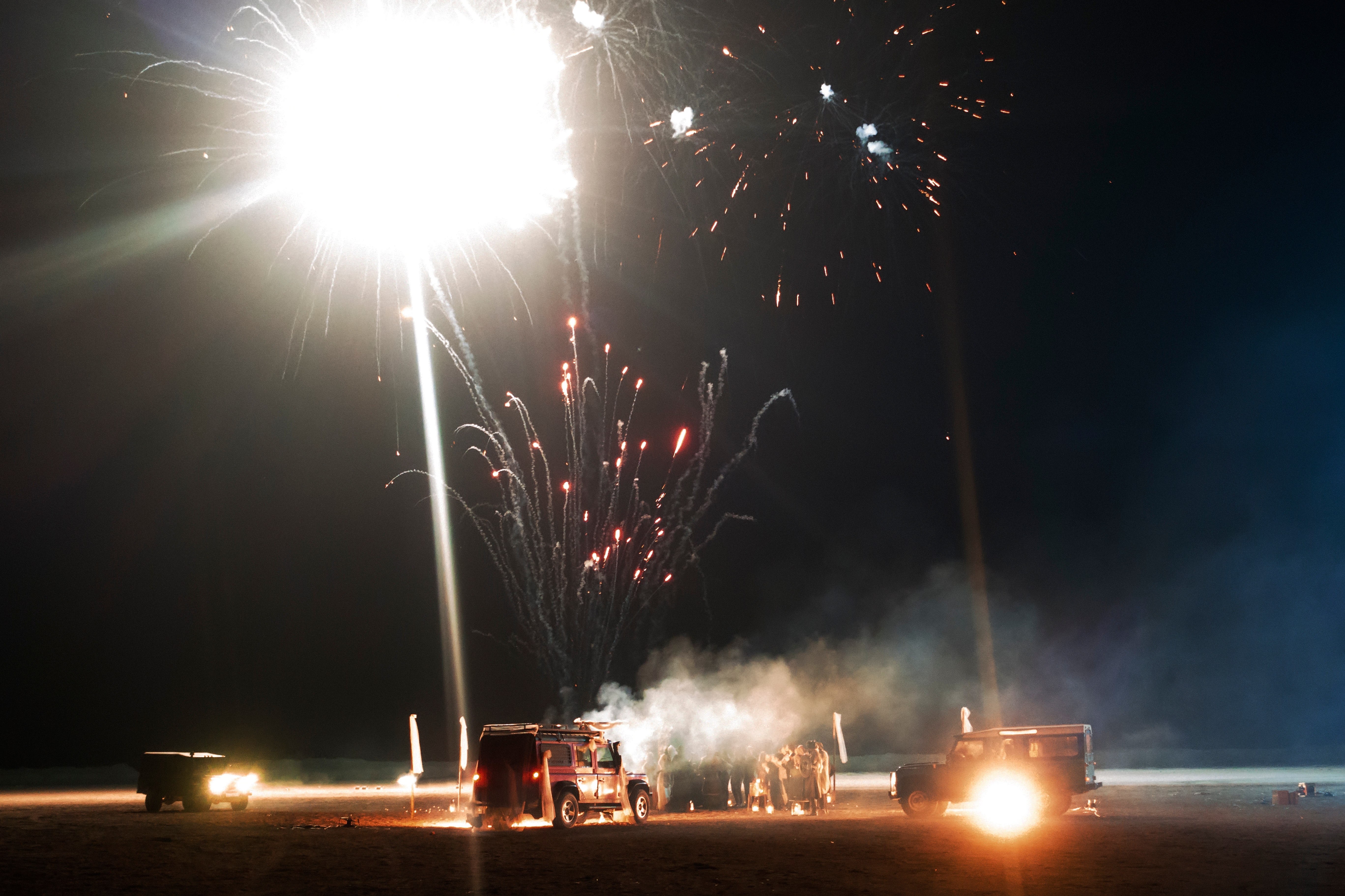 Fireworks light up the night sky above people gathered around vehicles, creating a festive atmosphere