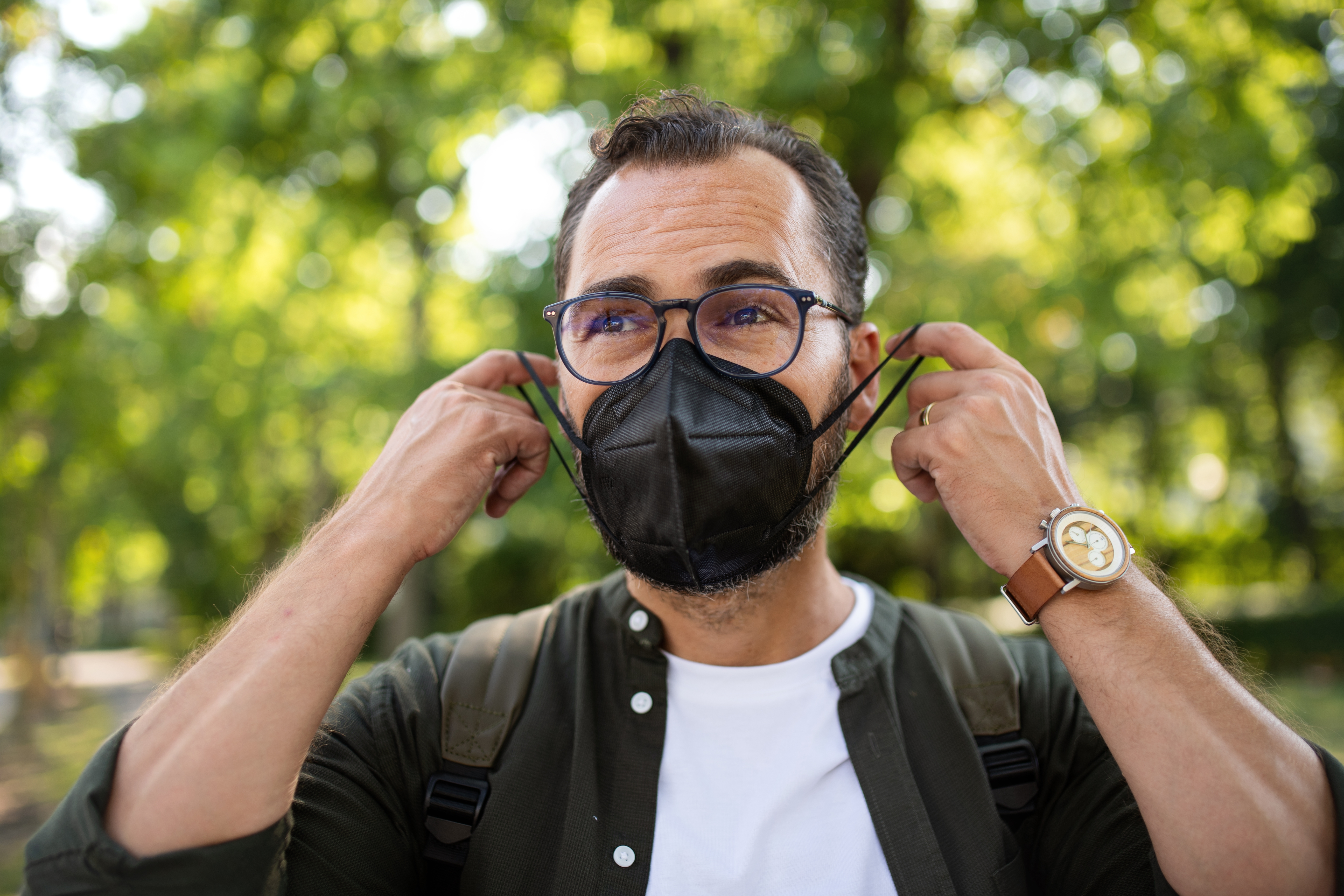 Person outdoors adjusting a black face mask, wearing glasses, a white shirt, and a casual jacket, set against a blurred greenery background