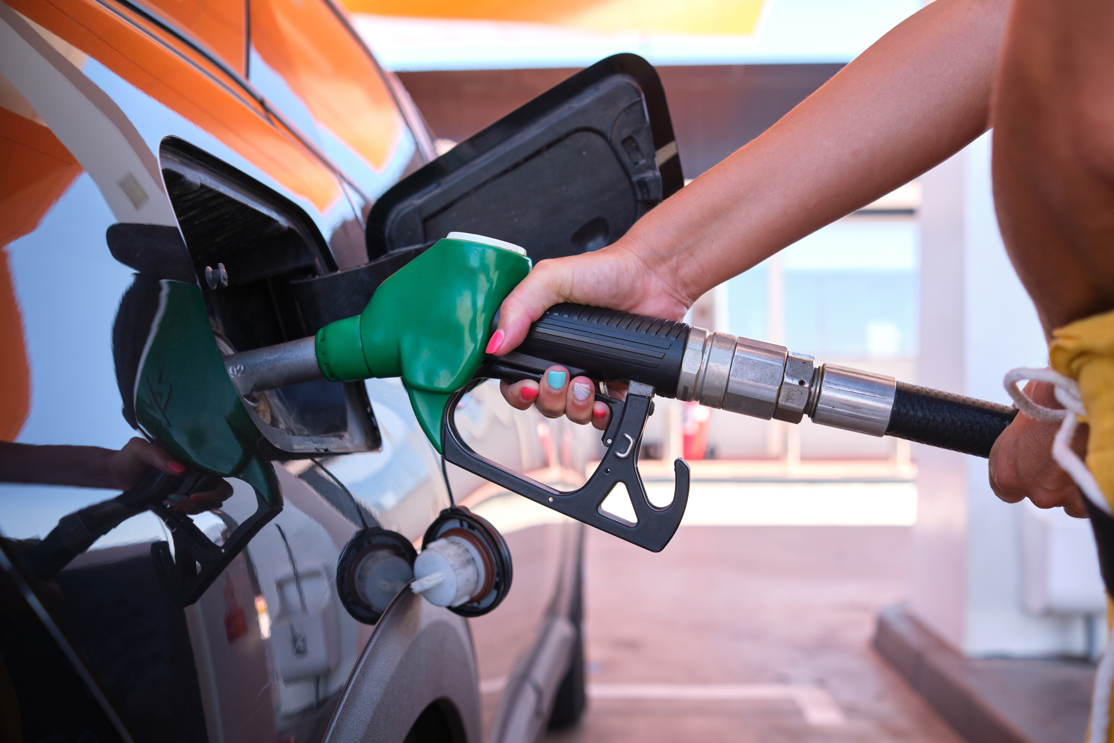 Person refuels car at gas station, holding a green fuel nozzle. The car's fuel tank door is open