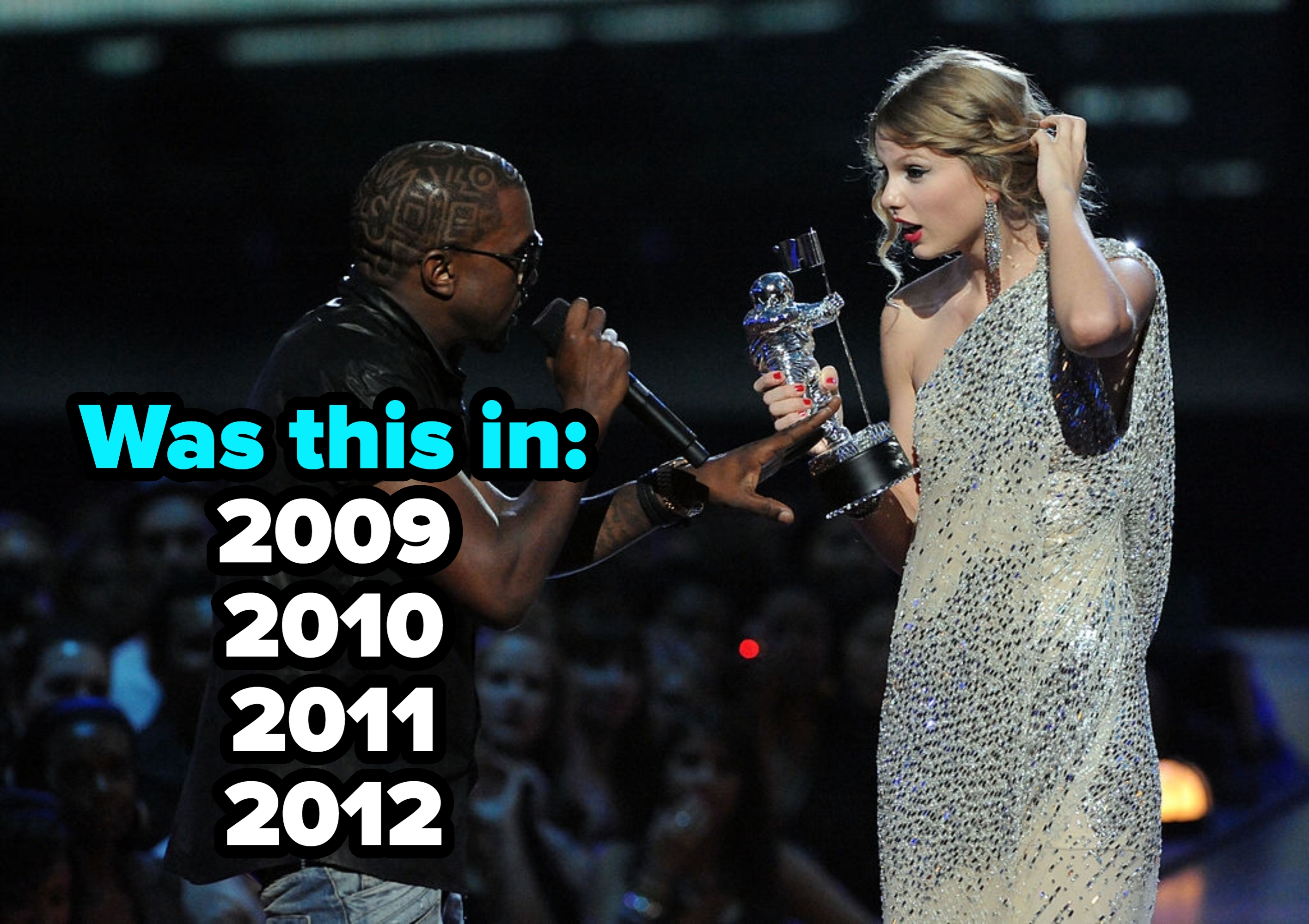 A musician interrupts an artist in a shimmering dress holding a trophy onstage during an award ceremony
