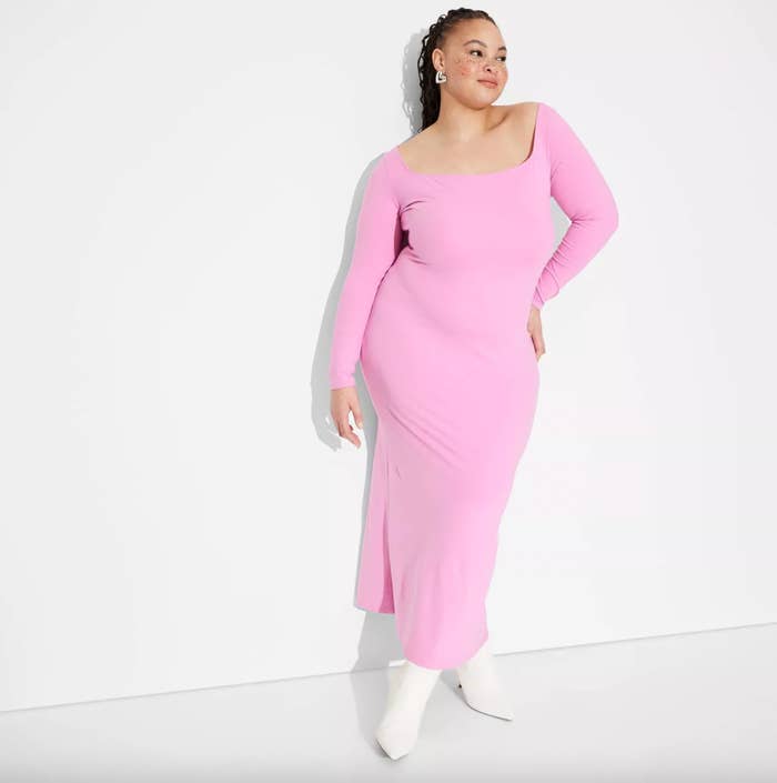 Person wearing a long, off-shoulder formal  poses confidently against a plain backdrop, adorned with driblet  earrings and achromatic  ankle boots