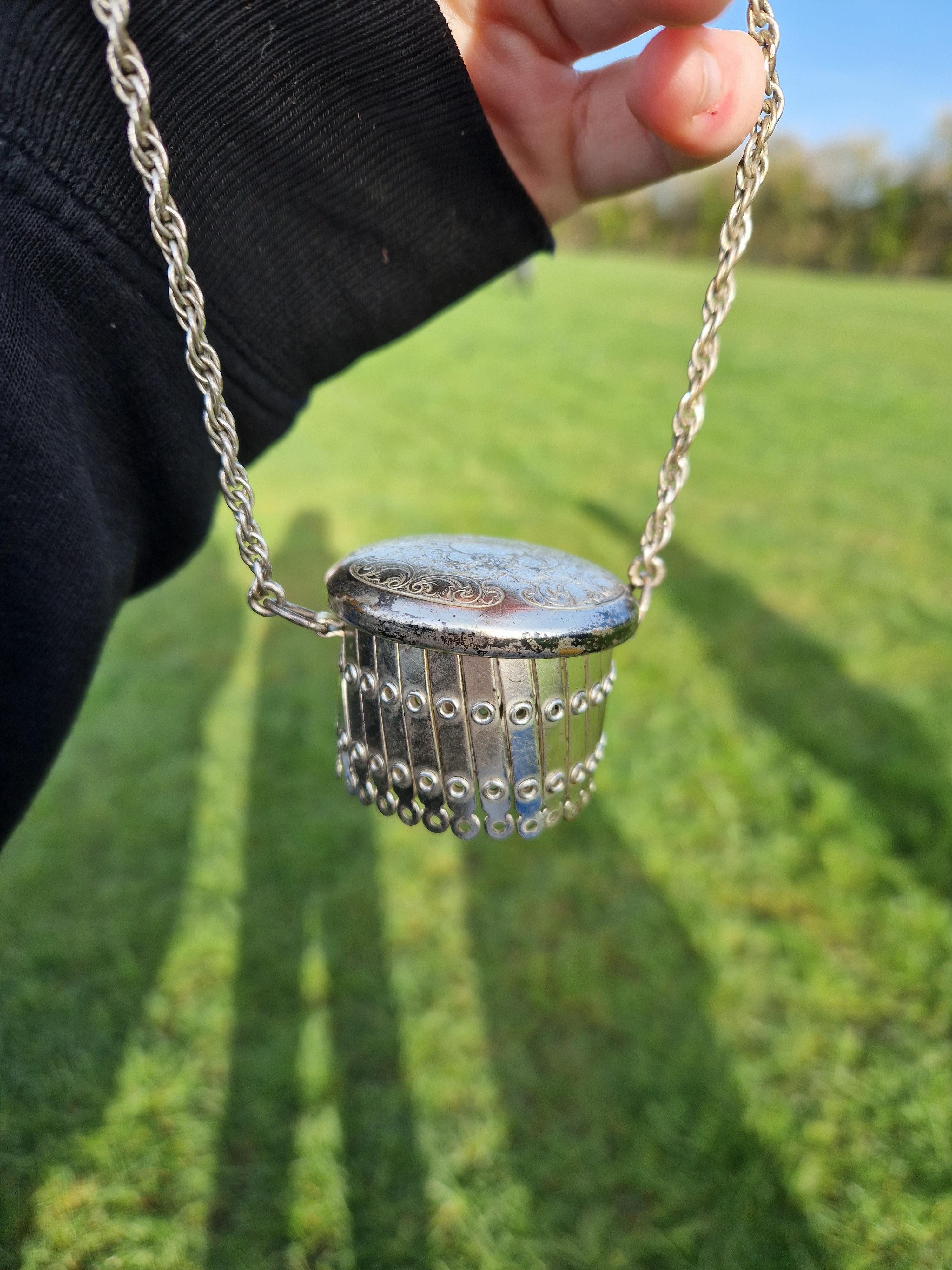 Person holds a chain with a larger metal section featuring a detailed design, against a grassy field backdrop