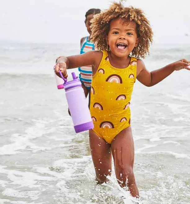 Anak di Rainbo Baju renang berpola W berjalan melalui gelombang dangkal, dengan senang hati memegang botol air ungu