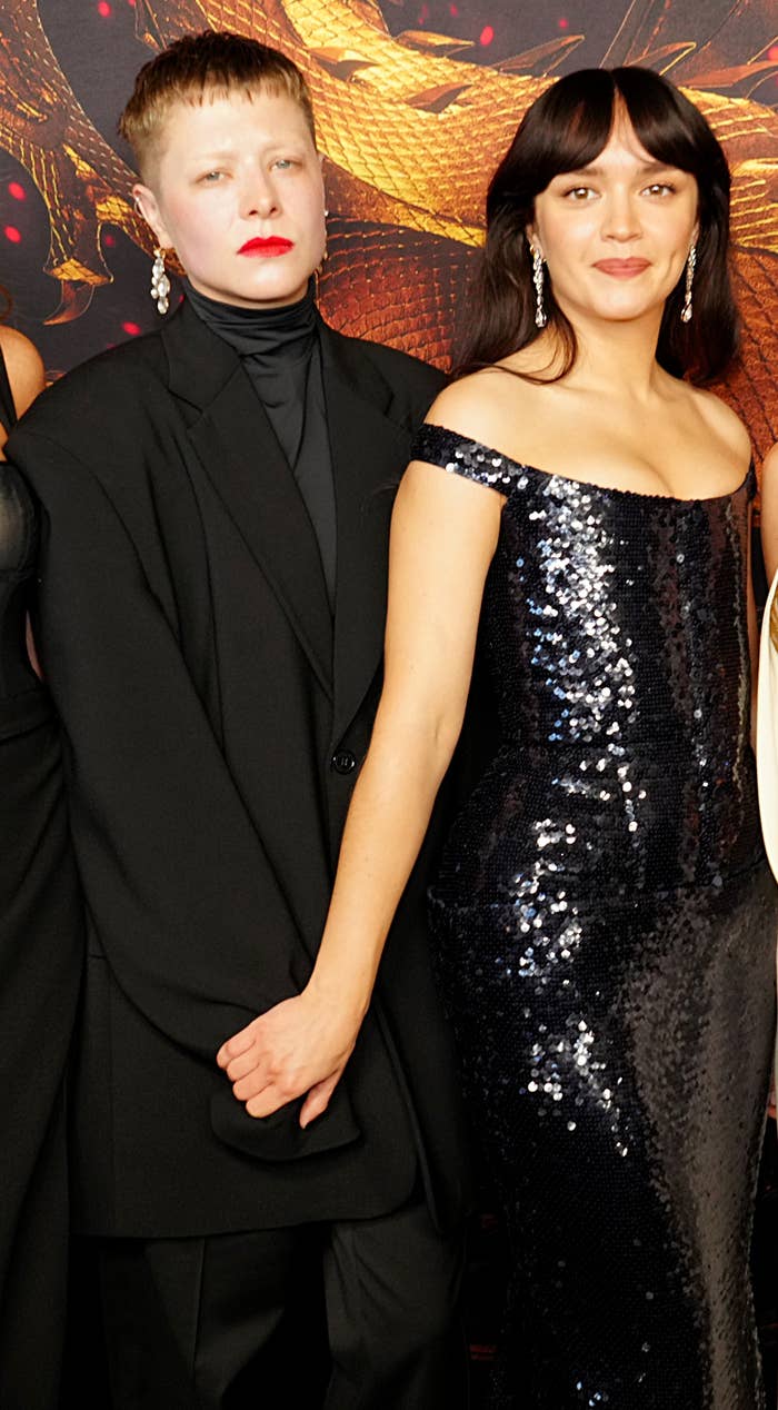 A group of people poses at an event. They wear a variety of stylish outfits, including a sequin dress, sheer and structured tops, and elegant suits