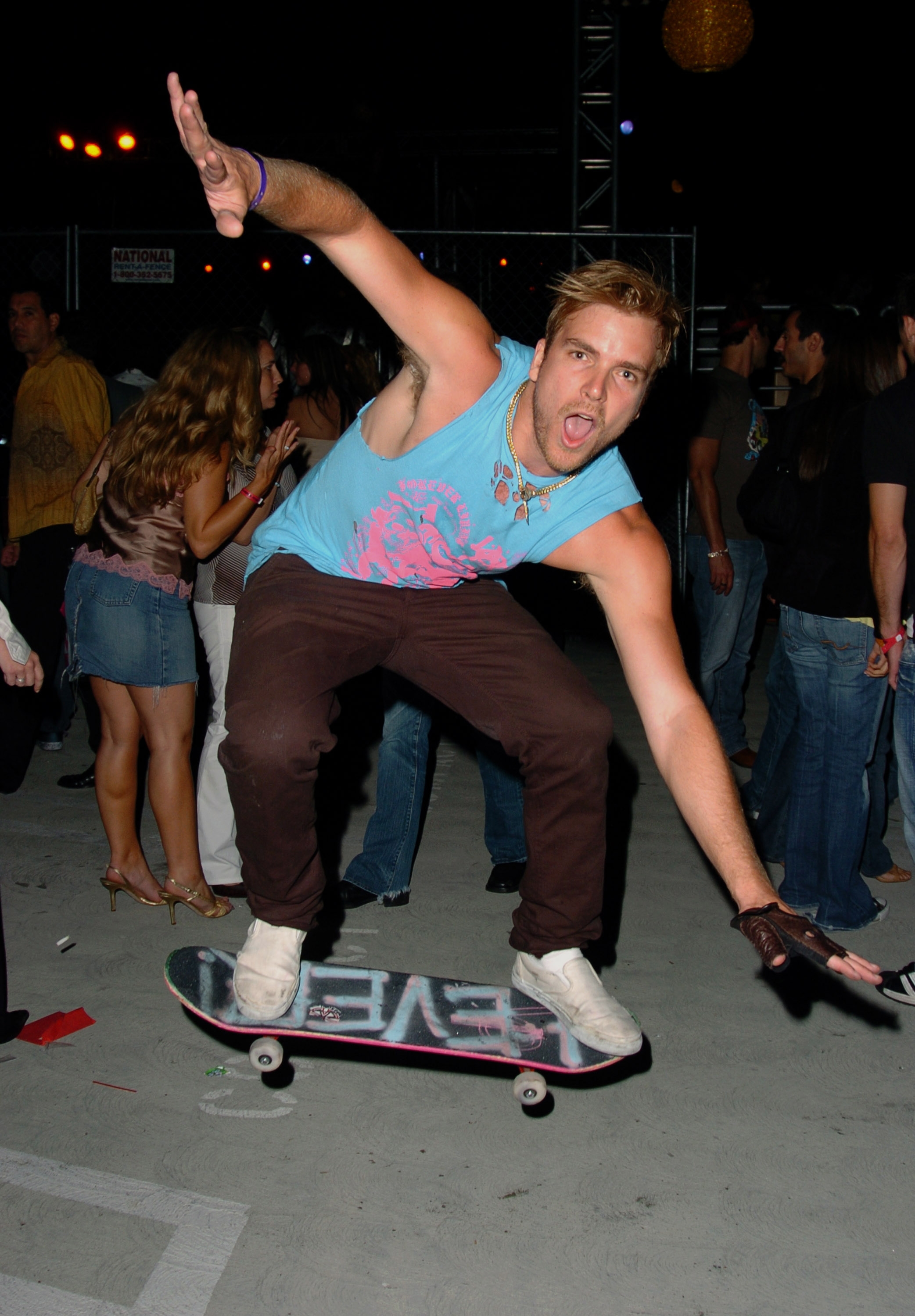 Person skateboarding mid-trick at a nighttime event, wearing a sleeveless graphic tee and brown pants, surrounded by casually dressed attendants
