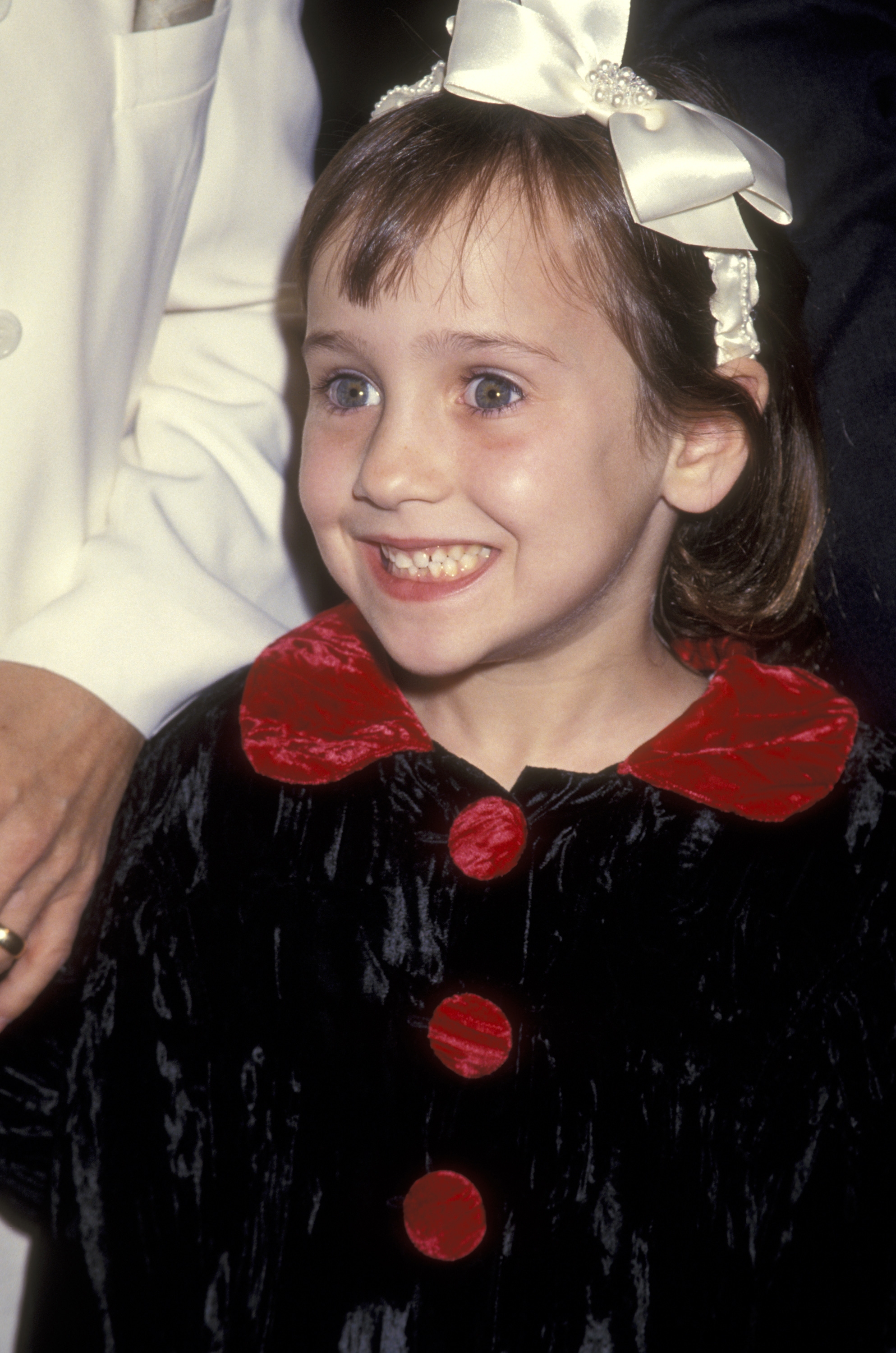 Child smiling broadly, wearing an elegant dress with large buttons and a decorative hairband
