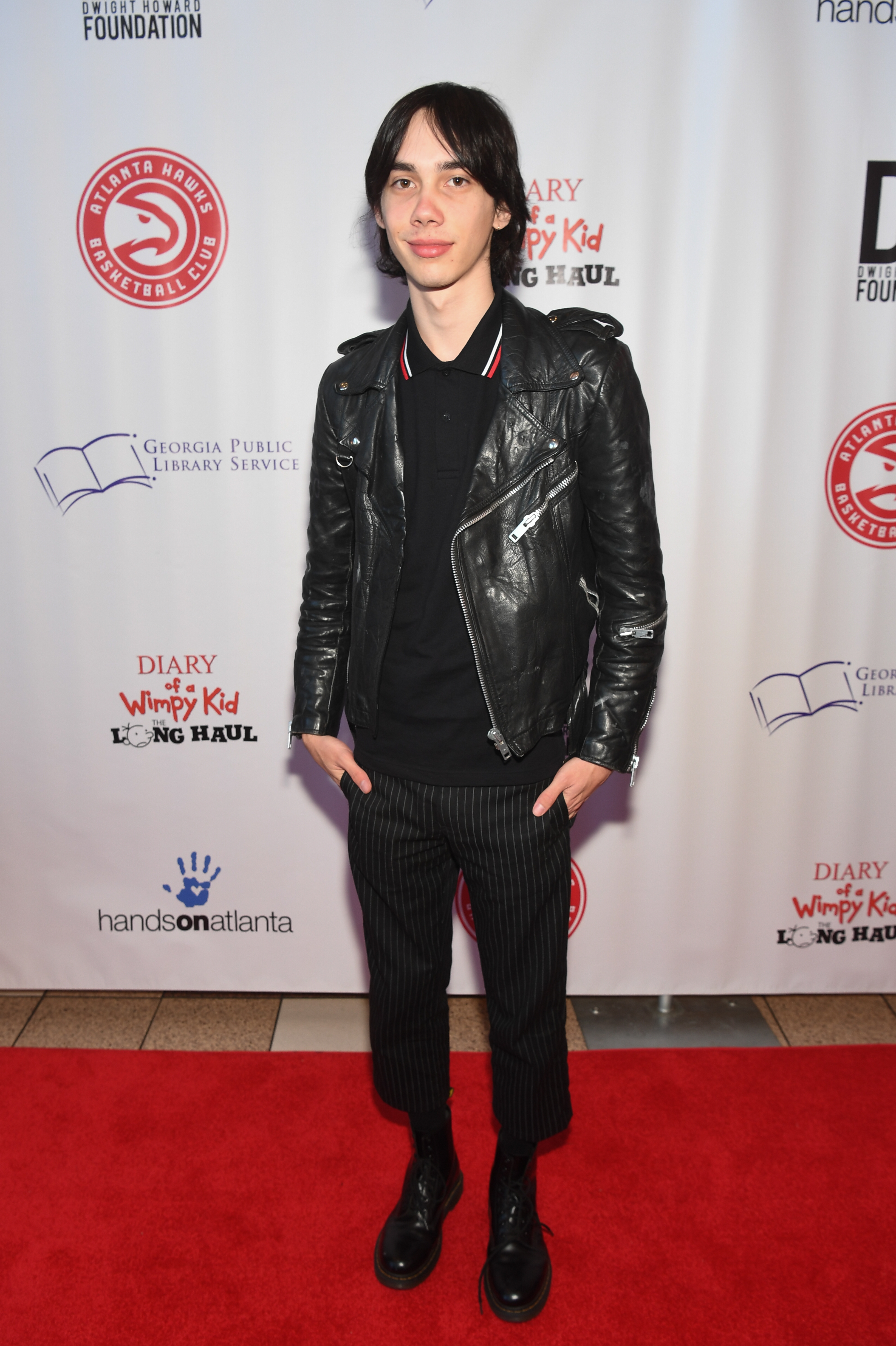 Person on red carpet wearing a black leather jacket, black shirt, striped pants, and black boots, at an event with "Diary of a Wimpy Kid" backdrop