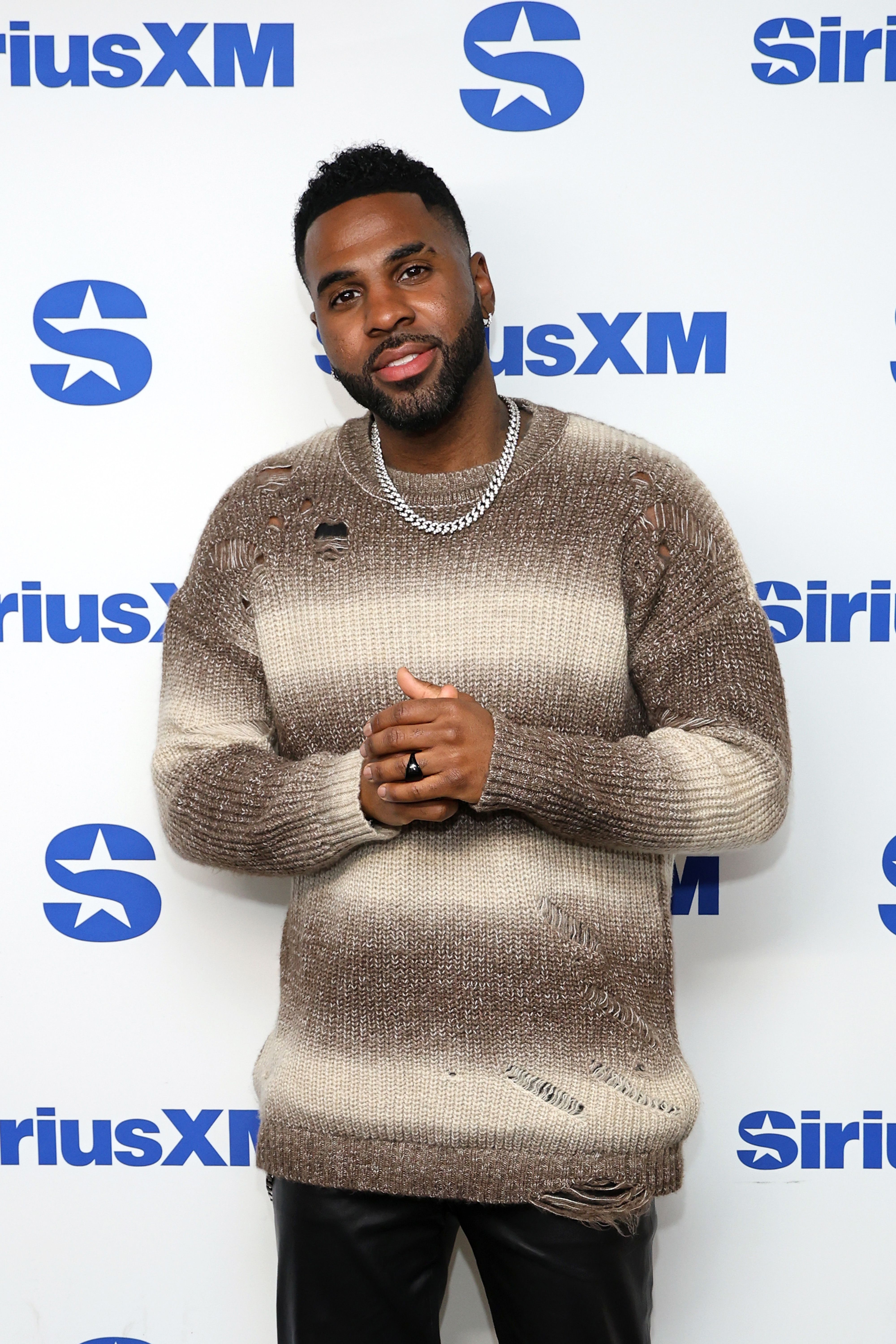 Person in a distressed knit sweater poses at a SiriusXM event backdrop, wearing a chain necklace