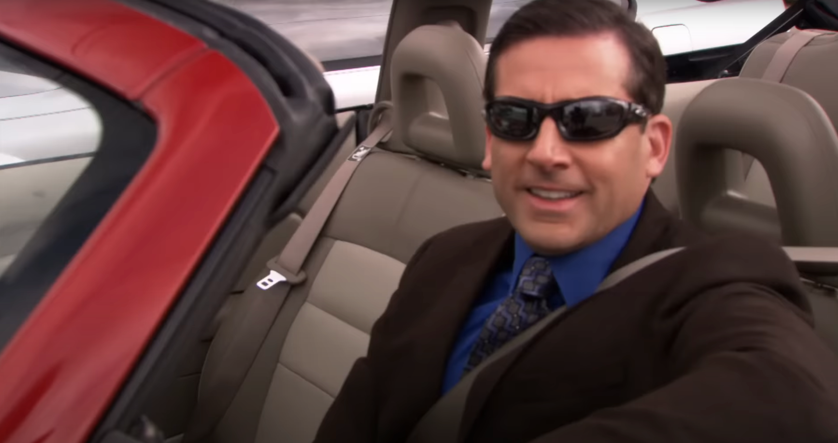 Michael Scott sits in a convertible, wearing a suit and tie, smiling at the camera