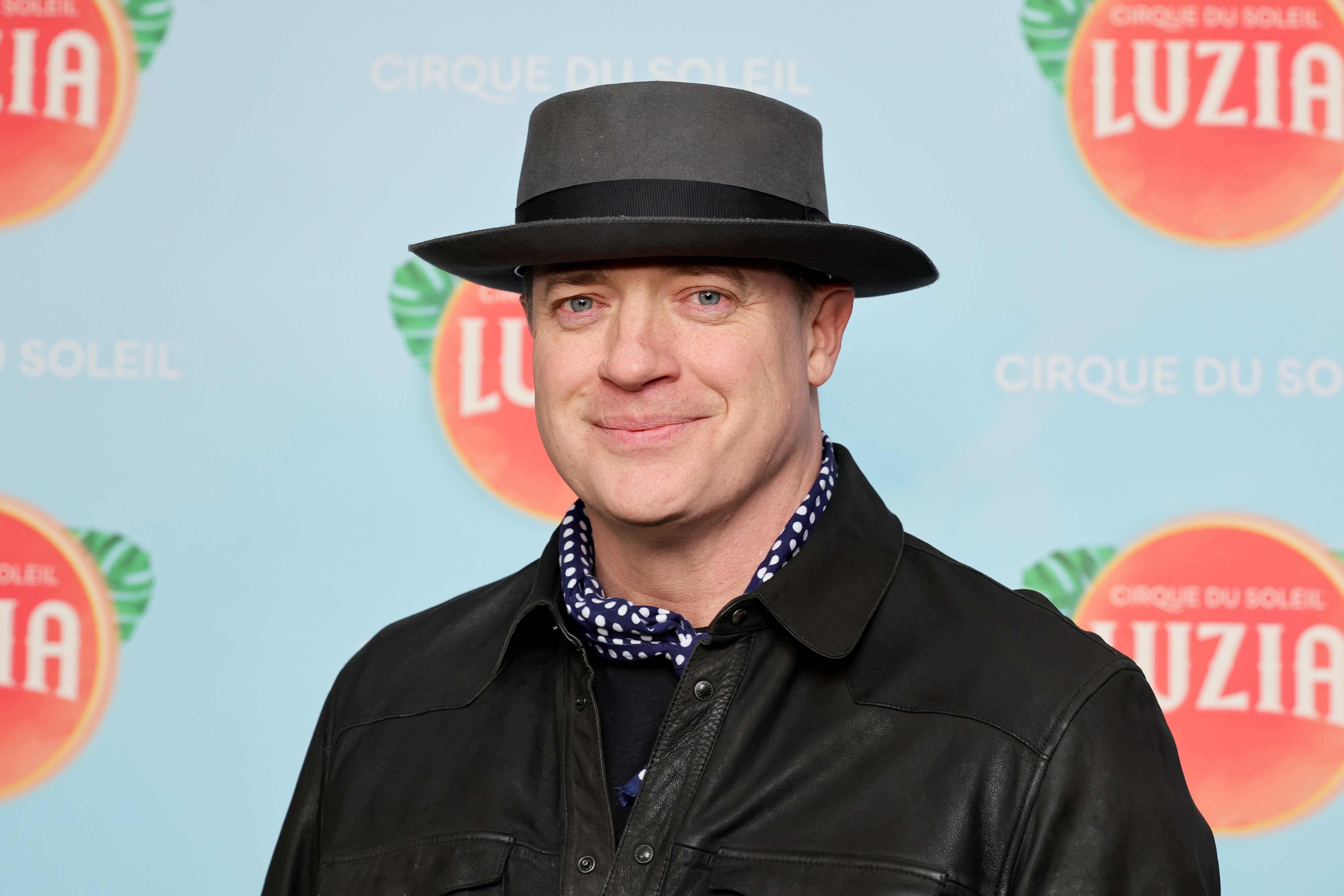 A person wearing a black jacket and hat, smiling at a &quot;Cirque du Soleil: Luzia&quot; event backdrop