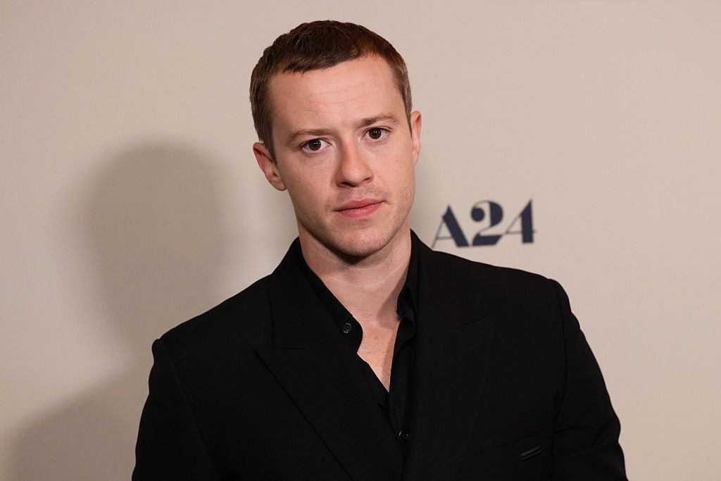 A person in formal attire stands against a backdrop with "A24" visible, often used for film-related events