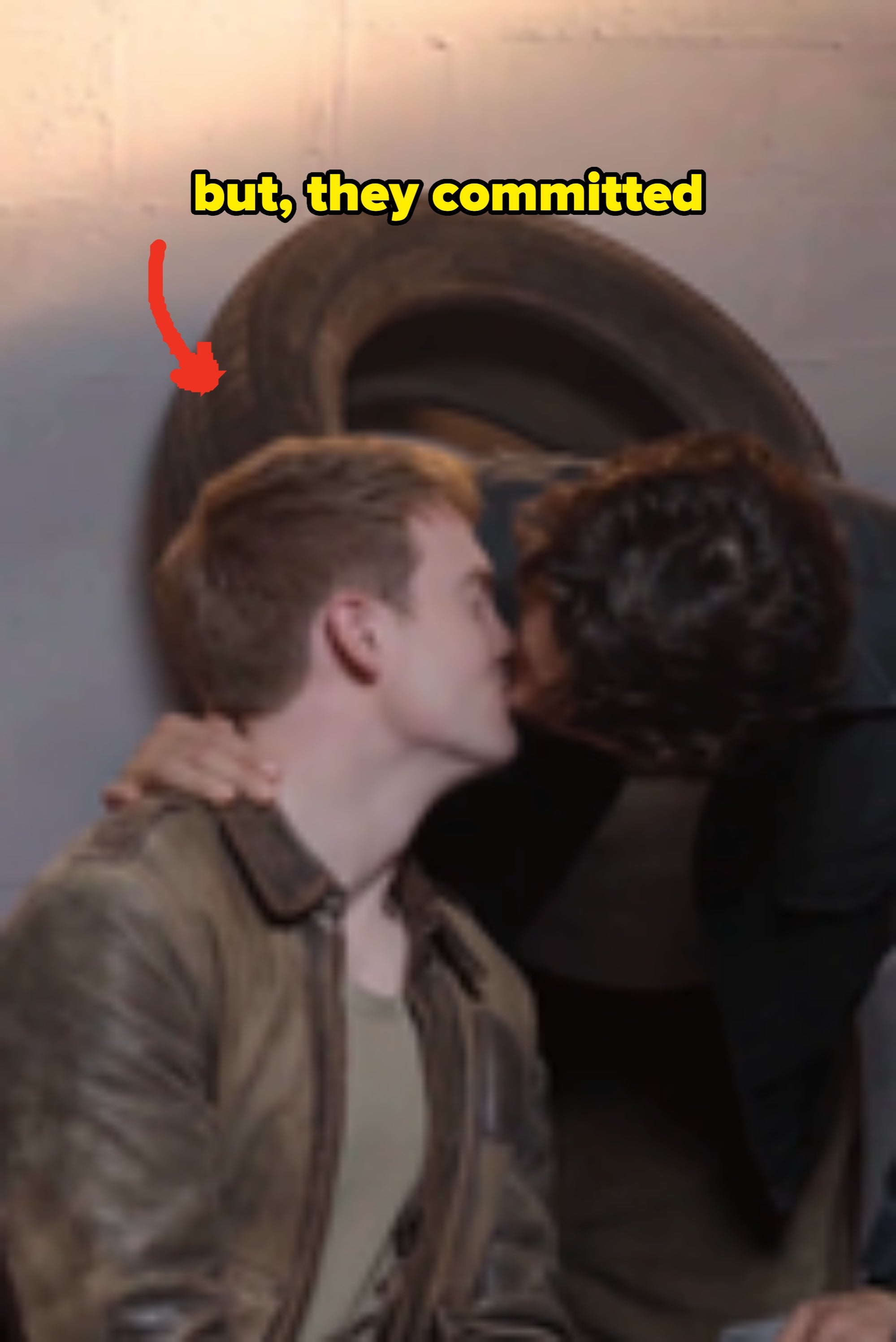Two individuals share a kiss; one wears a leather jacket, the other a dark top. The scene is casual, against an industrial backdrop