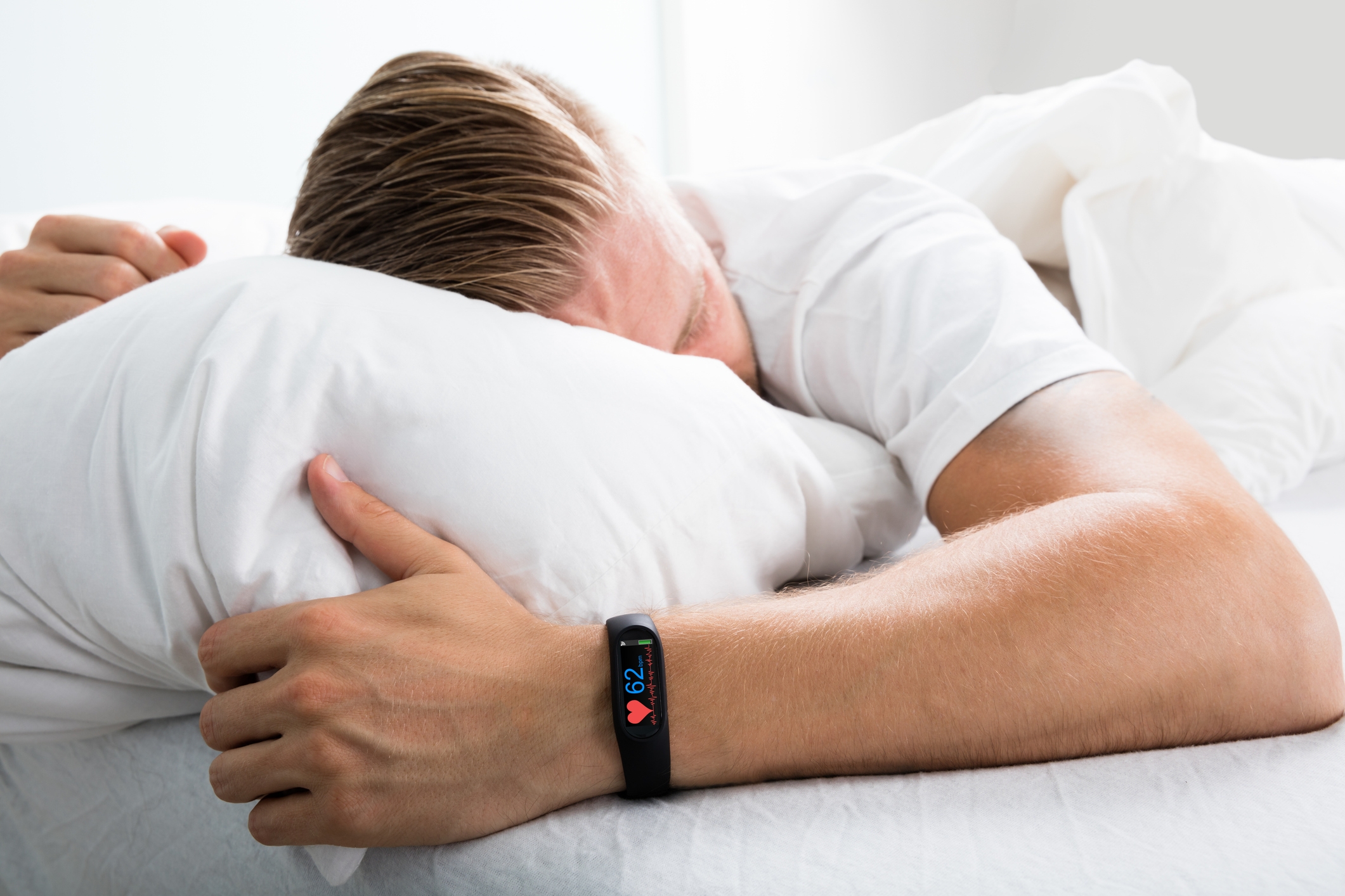 Personen som sover på en seng, iført en treningsspor som viser hjertefrekvens. De har en avslappet holdning med ansikt delvis skjult av en pute