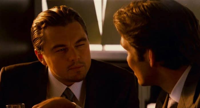 Two men in a serious conversation at a dimly lit table, one squinting slightly, wearing suits, creating an intense and focused atmosphere