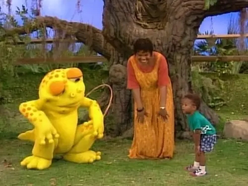 A woman and a child interact with a large, colorful frog costume character in a garden setting
