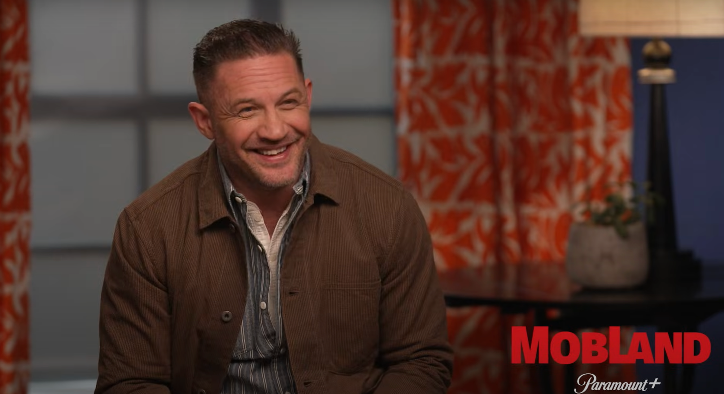 Smiling man in casual jacket sits in front of red-patterned curtains, “Mobland” and “Paramount+” logos at bottom right