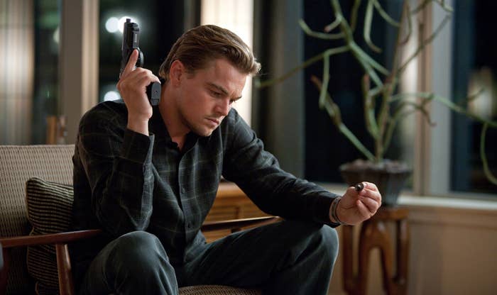 A person sits thoughtfully in a chair, holding a spinning top in one hand and a gun in the other, suggesting tension or decision-making