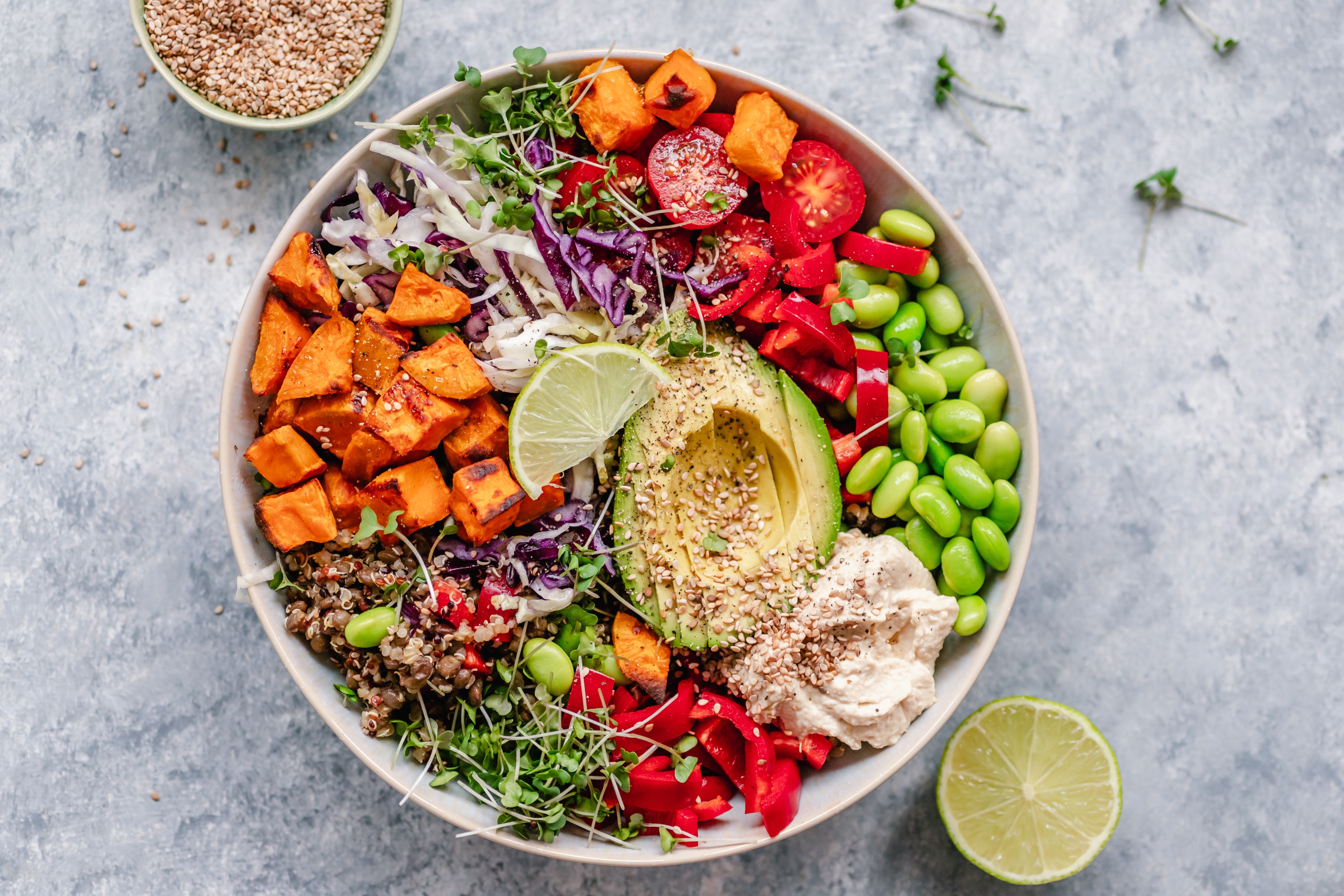 En pulserende skål med quinoa, stekte søtpoteter, avokado, edamame, rødkål, tomater, hummus og spirer, toppet med kalk og sesamfrø