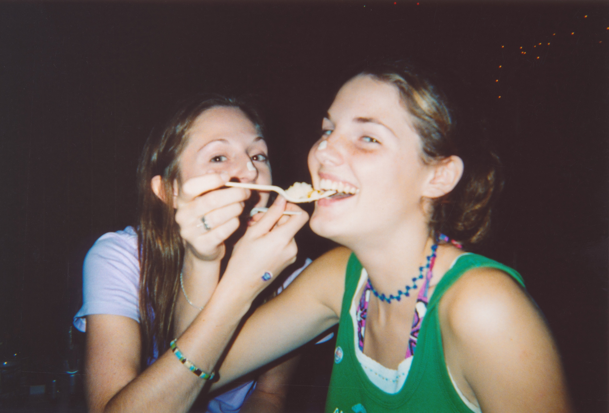 Two people laughing and sharing food with a fork, wearing casual tank tops and beaded jewelry. One feeds the other in a playful moment