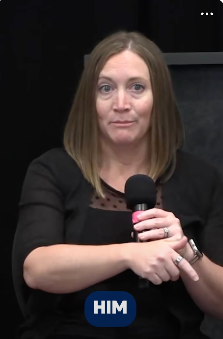 A person holds a microphone, pointing toward someone off-camera, wearing a black top with sheer details, seated indoors, engaged in a discussion