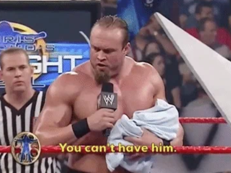 Wrestler in a ring holds a baby covered with a blanket, saying "You can't have him," during a wrestling event. An official stands nearby