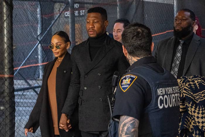 A couple holds hands outside a New York State courthouse, flanked by security and individuals in formal attire