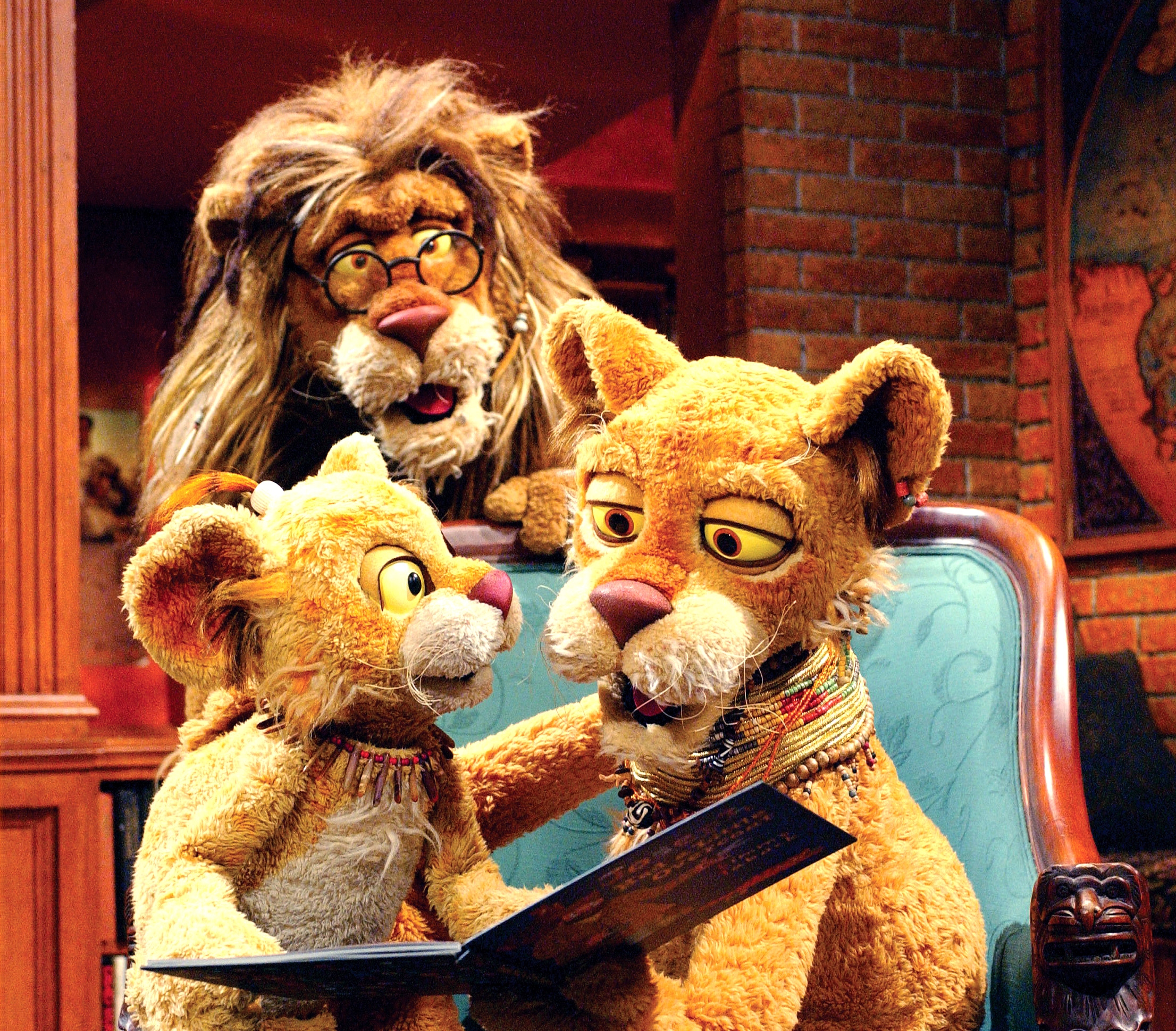Three lion puppet characters, including an adult and a cub, read a book together in a lively setting