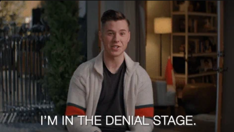 A person sits indoors, smiling, with text overlay reading, "I'M IN THE DENIAL STAGE."