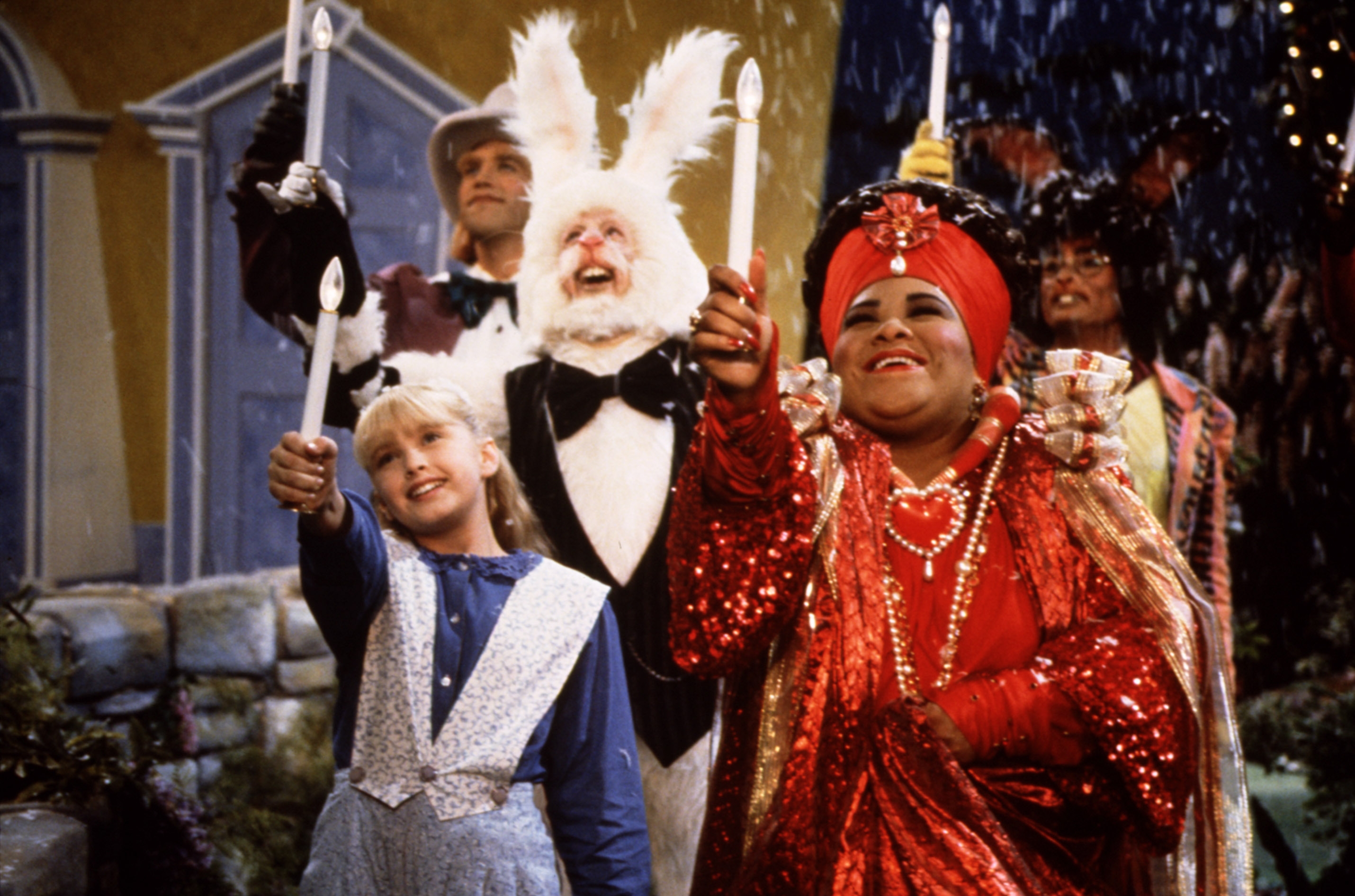 Group of people in theatrical costumes holding candles; one dressed as a white rabbit and another in ornate red, suggesting a festive scene