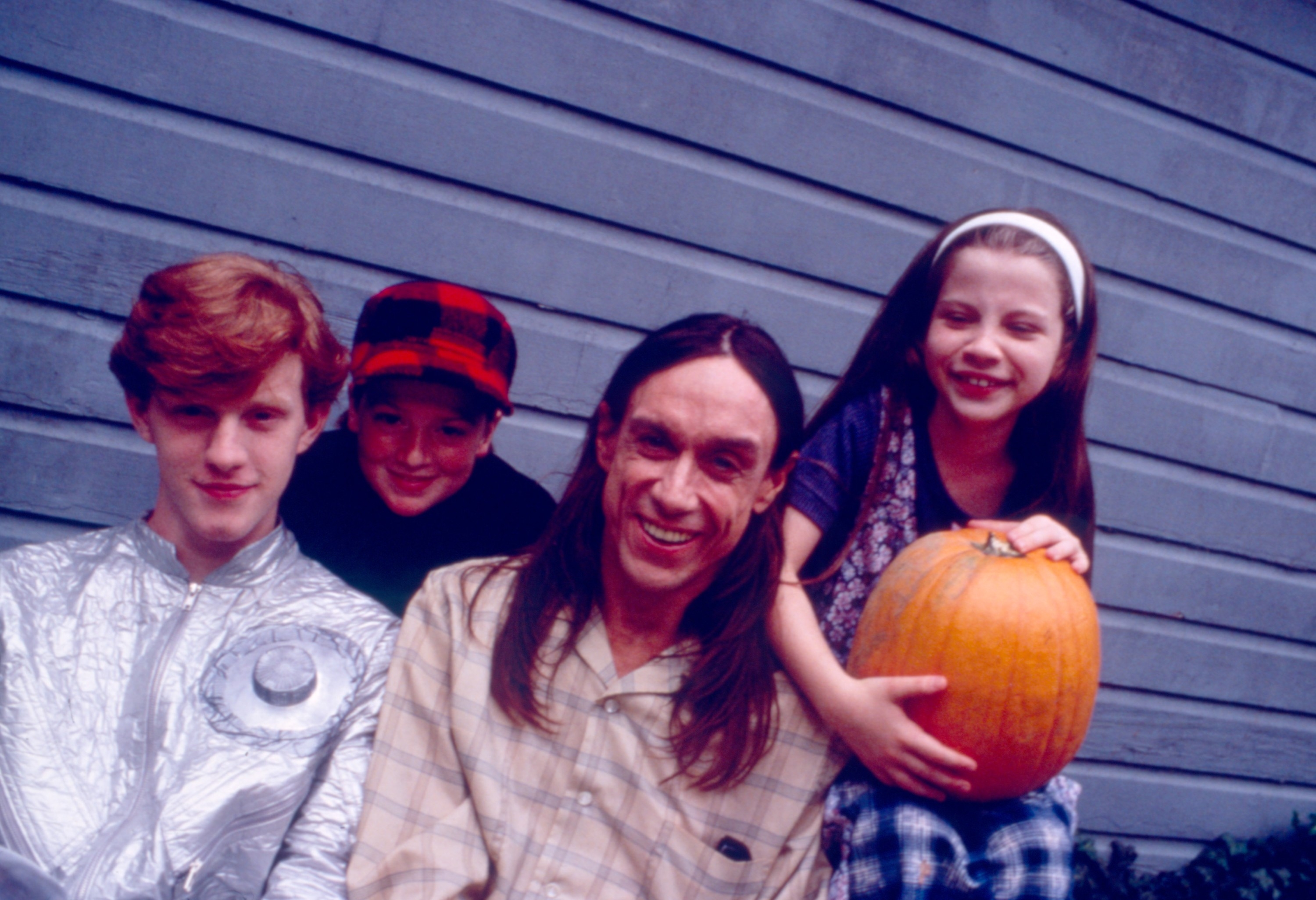 A group photo of three kids and an adult smiling. One child holds a pumpkin. Everyone is casually dressed, sitting in front of a wooden wall