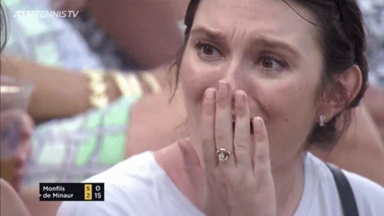 A woman looks emotional, covering her mouth with her hand, during a tennis match. Score display in the corner shows Monfils vs. de Minaur