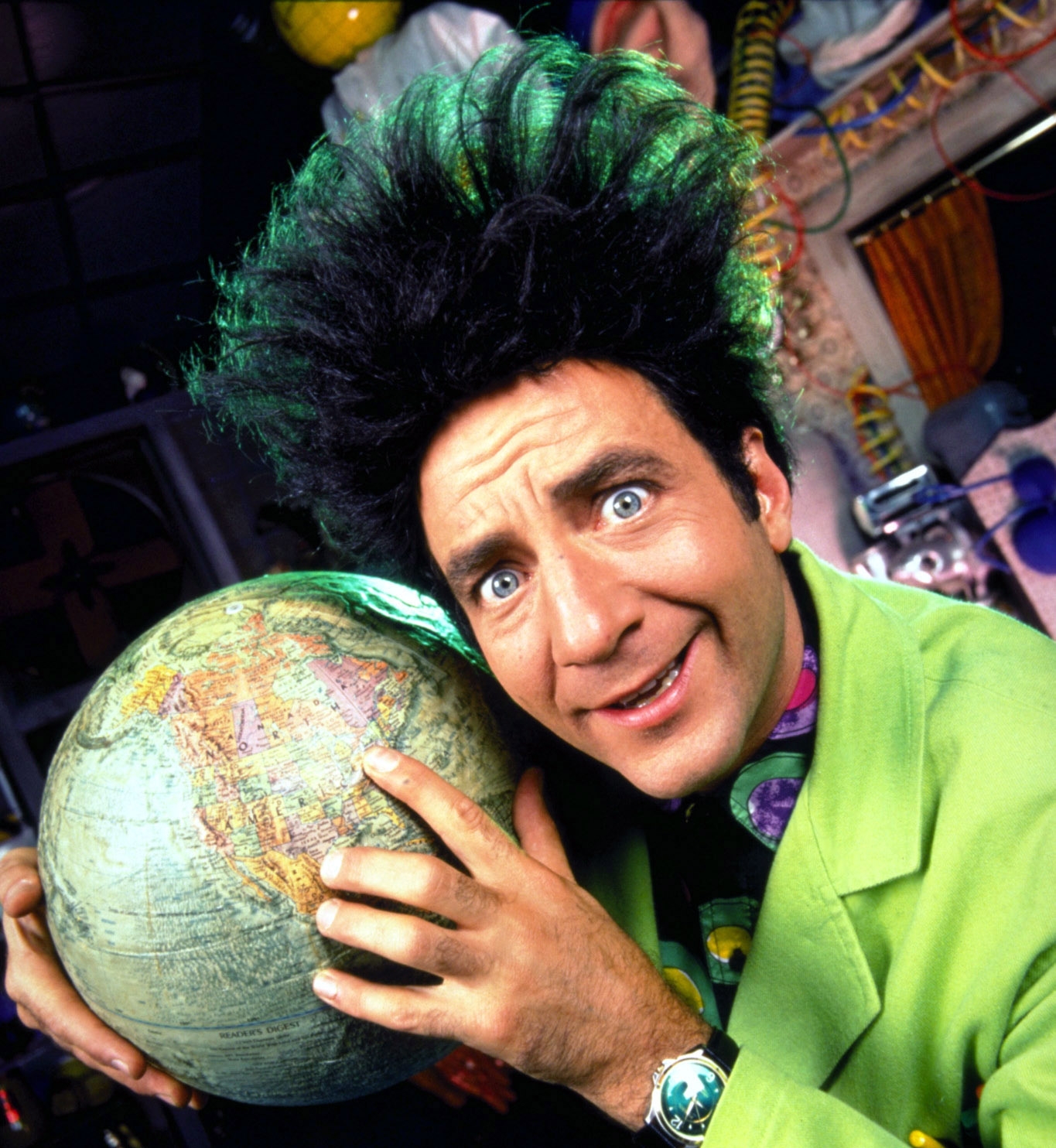 Person with wild hair holds a globe, wearing a quirky lime green suit and patterned shirt, smiling energetically in a playful pose