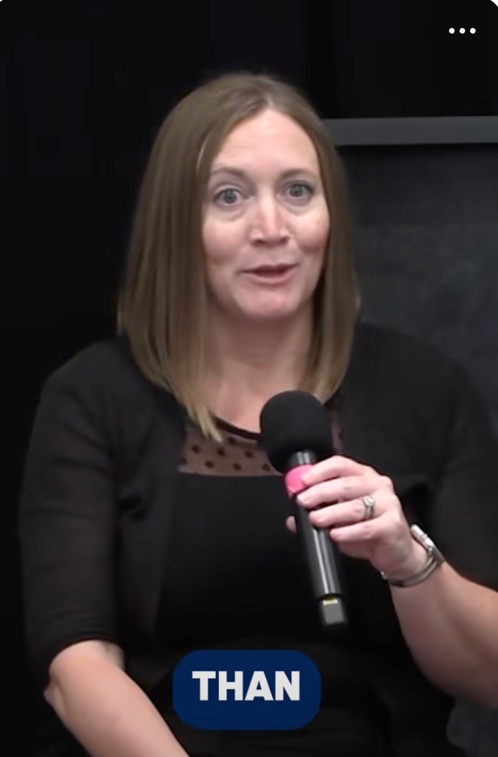 A person with shoulder-length hair holds a microphone, speaking in a seated position